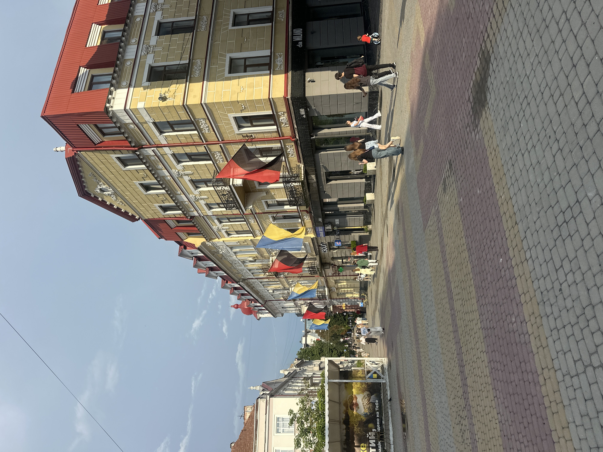 A street scene in Ternopil, Ukraine, captures a pedestrian promenade lined with ornate yellow buildings adorned with red roofs and balconies. Ukrainian flags are prominently displayed along the facades, some of them partially obscured by building architecture. A diverse group of pedestrians strolls along the paved walkway, which is laid with interlocking rectangular stones in shades of yellow, pink, and gray. The atmosphere is relaxed, with individuals walking, conversing, and children playing. A young child is seen on a small ride-on toy. The sky above is a mixture of blue and gray clouds, suggesting dynamic weather conditions. The composition of the image is a medium shot, looking down a street.