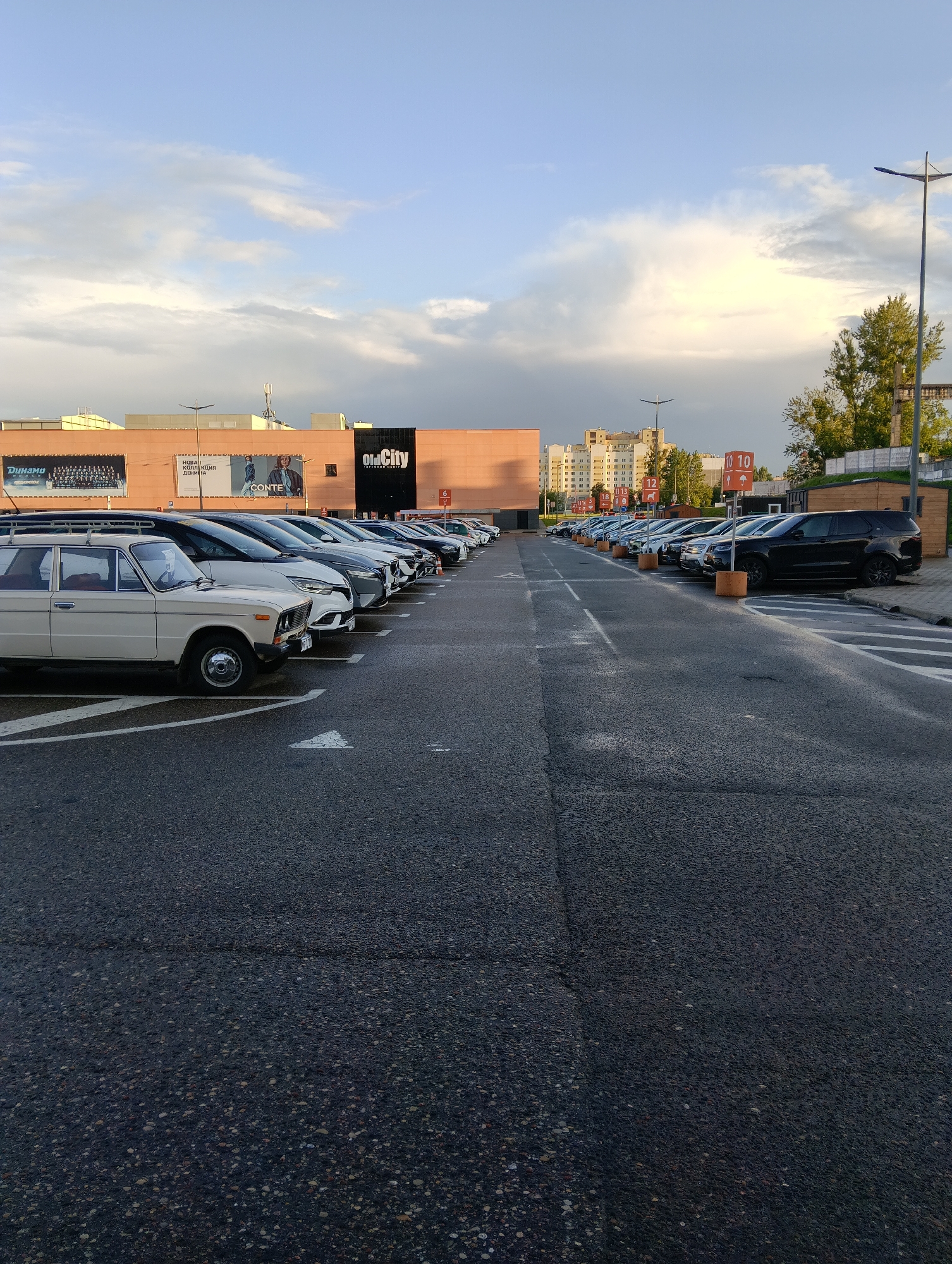 An outdoor parking lot scene in Hrodna, Belarus. In the foreground, a row of parked cars stretches into the distance. The closest car is a light-colored sedan with a roof rack. Behind it are several other vehicles, including SUVs and sedans of various colors. The parking lot pavement is asphalt, with white directional arrows painted on the ground. In the background, a large, modern commercial building with an orange facade is visible. The building has the "Old City" logo displayed prominently. Several trees and streetlights are also present in the scene. The sky above is partly cloudy with a mix of blue sky and white clouds.