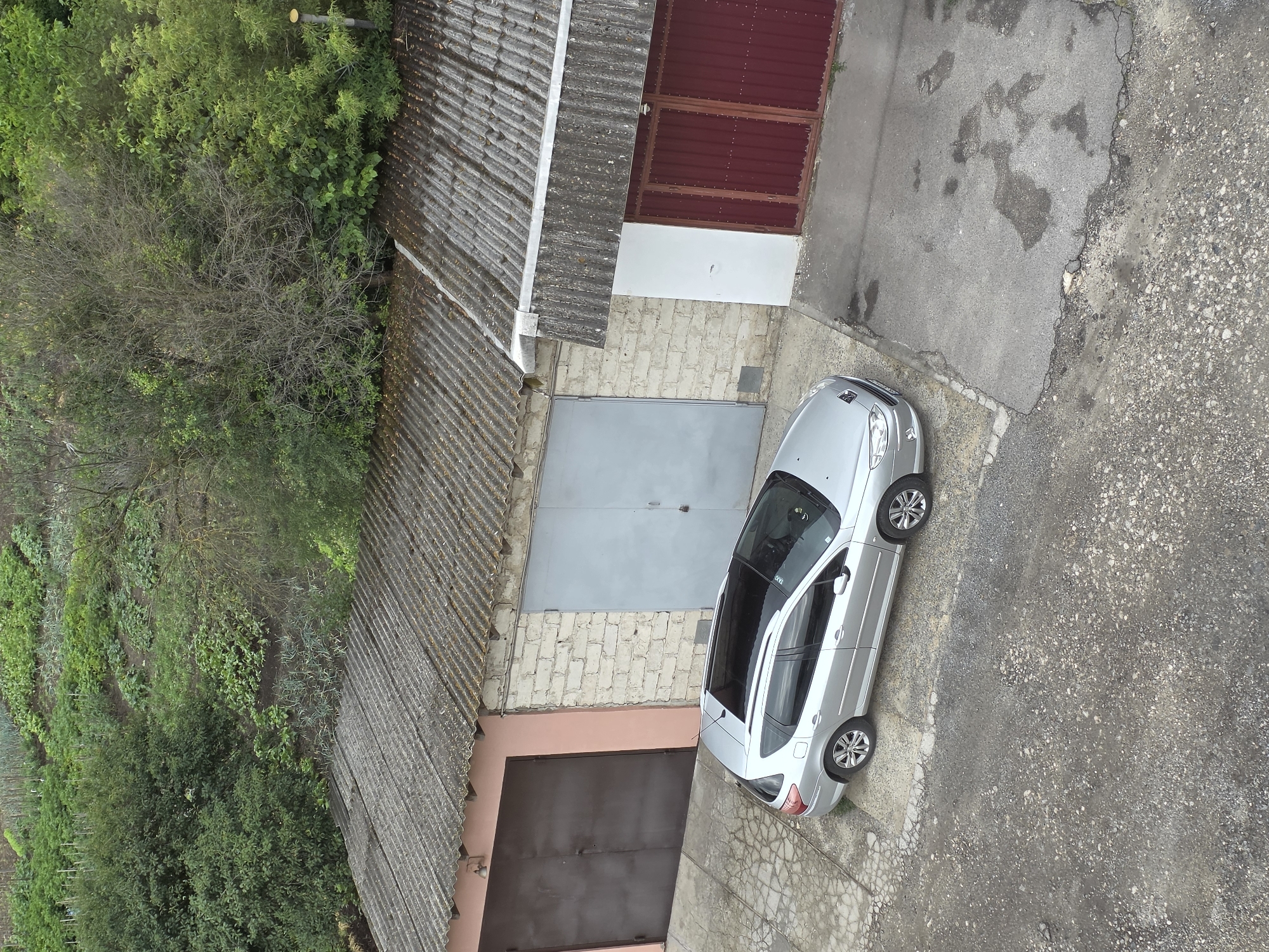 The image depicts a silver Peugeot estate car parked on a gravel and asphalt driveway in front of several garages. The garages have corrugated roofs and different colored doors: one is dark brown, another is light gray, and a third has dark red double doors. Behind the garages, a hillside is visible with lush green vegetation and what appears to be a cultivated garden or field. The scene is set in the city of Dubno, Ukraine. There are no people visible in the image.