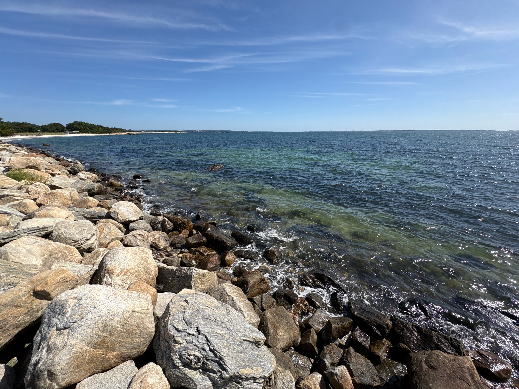 A rocky shoreline lines the edge of a body of water under a bright blue sky with thin, wispy clouds. The water is a deep blue, transitioning to a lighter, clear green closer to the shore, revealing the rocky seabed beneath. Waves gently lap against the stones. In the distance, a sandy beach is visible, bordered by a line of green trees. Further out, several structures and what appear to be ships or boats can be seen on the horizon. The foreground is dominated by large, weathered rocks of varying sizes and colors, some smooth and rounded, others rough and jagged. The scene is peaceful and natural, suggesting a coastal environment.