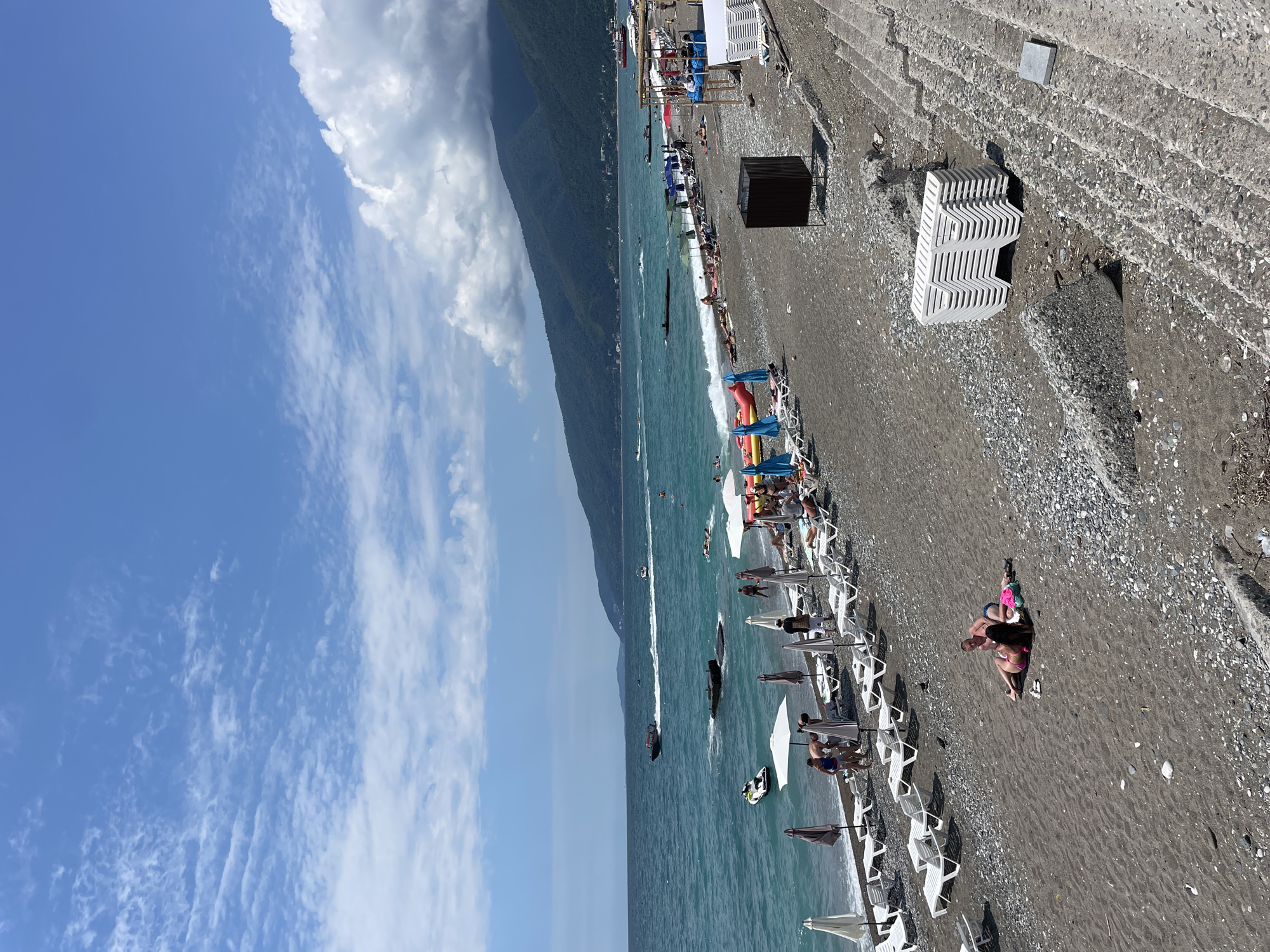 The image depicts a crowded beach scene in Gagra, Abkhazia, on a sunny day with scattered clouds. The beach is composed of dark sand and pebbles, with a line of white lounge chairs and umbrellas set up for sunbathers. In the turquoise water, several jet skis and boats are visible, along with people swimming and enjoying various water activities. A long, tree-covered mountain range forms the backdrop to the scene. The foreground features a concrete staircase leading down to the beach. Several people are scattered across the beach, some lounging on towels and others walking. The overall atmosphere is one of leisure and summer recreation.