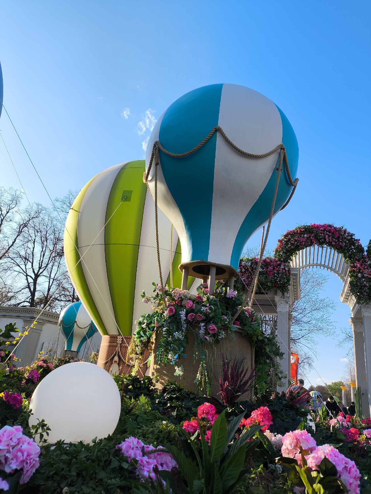 The image shows a bright, sunny outdoor scene with whimsical, oversized decorative hot air balloons nestled among flower beds. Two prominent balloons are striped blue and white and green and white, respectively. The balloons' baskets are adorned with vibrant arrangements of pink and white roses and cascading greenery.
A white, spherical lamp sits within the flower bed, adding to the fanciful atmosphere. In the background, a white arched structure is decorated with more flowers, creating a garden-like setting.
The image was taken at the Victory Park on Poklonnaya Hill in Moscow, Russia. People can be seen walking in the background.