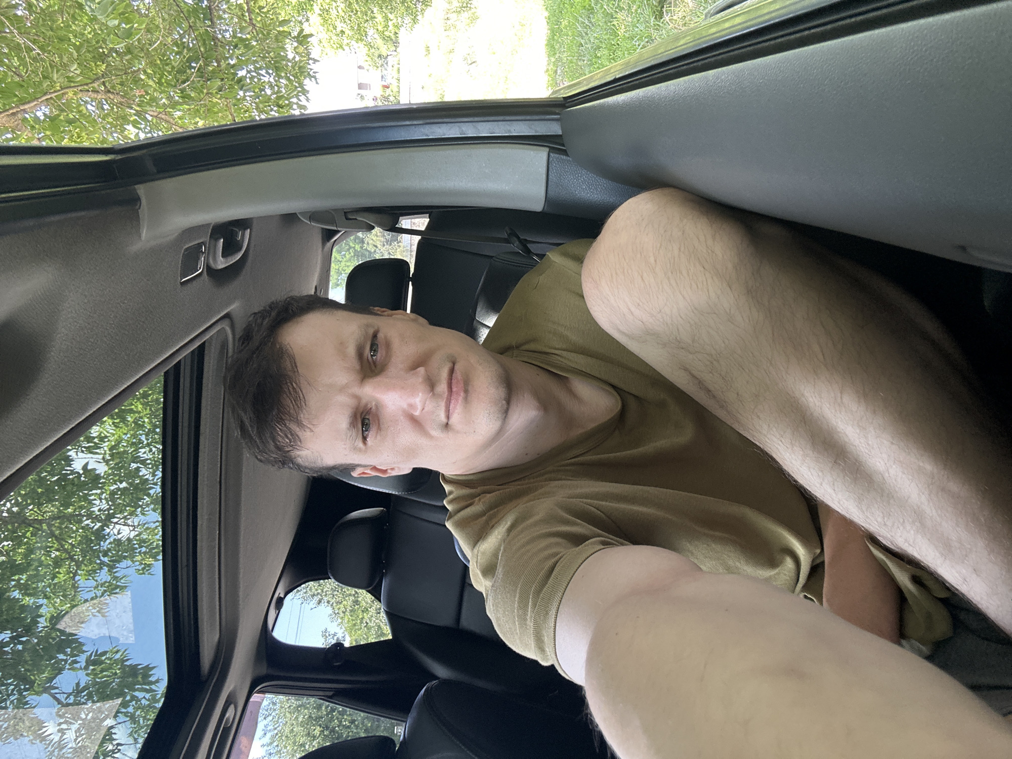 In the original image, a man is captured in a selfie taken from the back seat of a car. He is wearing a tan t-shirt and has short brown hair. His gaze is directed towards the camera with a neutral expression. The car's interior is visible, including black leather seats and a sunroof that reveals green trees and a bright sky, suggesting it's daytime with fair weather. The background outside the car shows a grassy area and trees, with hints of a building in the distance. The photo is oriented vertically, with the man's legs prominently in the foreground, partially obscuring the view of the car's interior. The scene is set in Makiv, Ukraine. No visible text is present in the image.