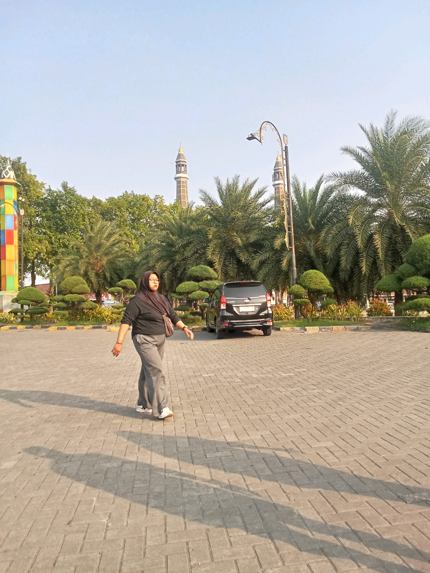 The image depicts a daytime scene in front of a mosque in Kaliwungu, Indonesia, characterized by paved ground and palm trees. A woman, wearing a dark hijab and gray pants, is walking in the foreground, casting a long shadow on the patterned pavement. Behind her, a black car is parked. In the background, the distinctive minarets of the mosque rise above lush green foliage, with a decorative, colorful pillar on the left side of the frame and a streetlamp to the right. The sky is clear and blue.