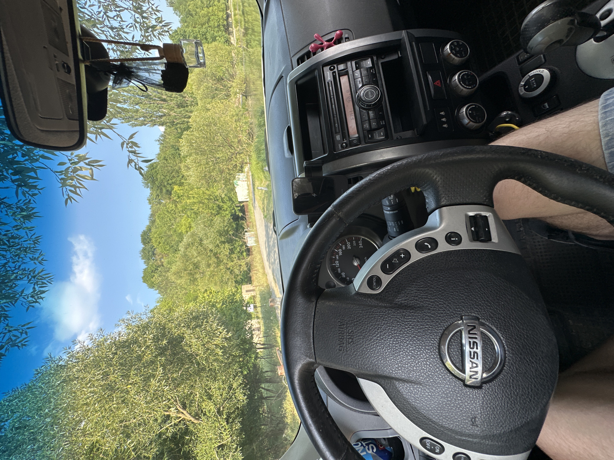 The image, taken from the interior of a Nissan vehicle, shows a view through the windshield of a scenic outdoor environment. The steering wheel and dashboard are prominently featured in the foreground. Beyond the car's interior, a road winds through a landscape characterized by lush green trees and foliage, with a body of water visible in the distance. The sky is clear blue with some scattered clouds, suggesting a sunny day. The location is identified as Makiv, Ukraine. There are no people visible within the car, but the perspective suggests the driver's legs are present. The dashboard includes the car's stereo system and various controls.
