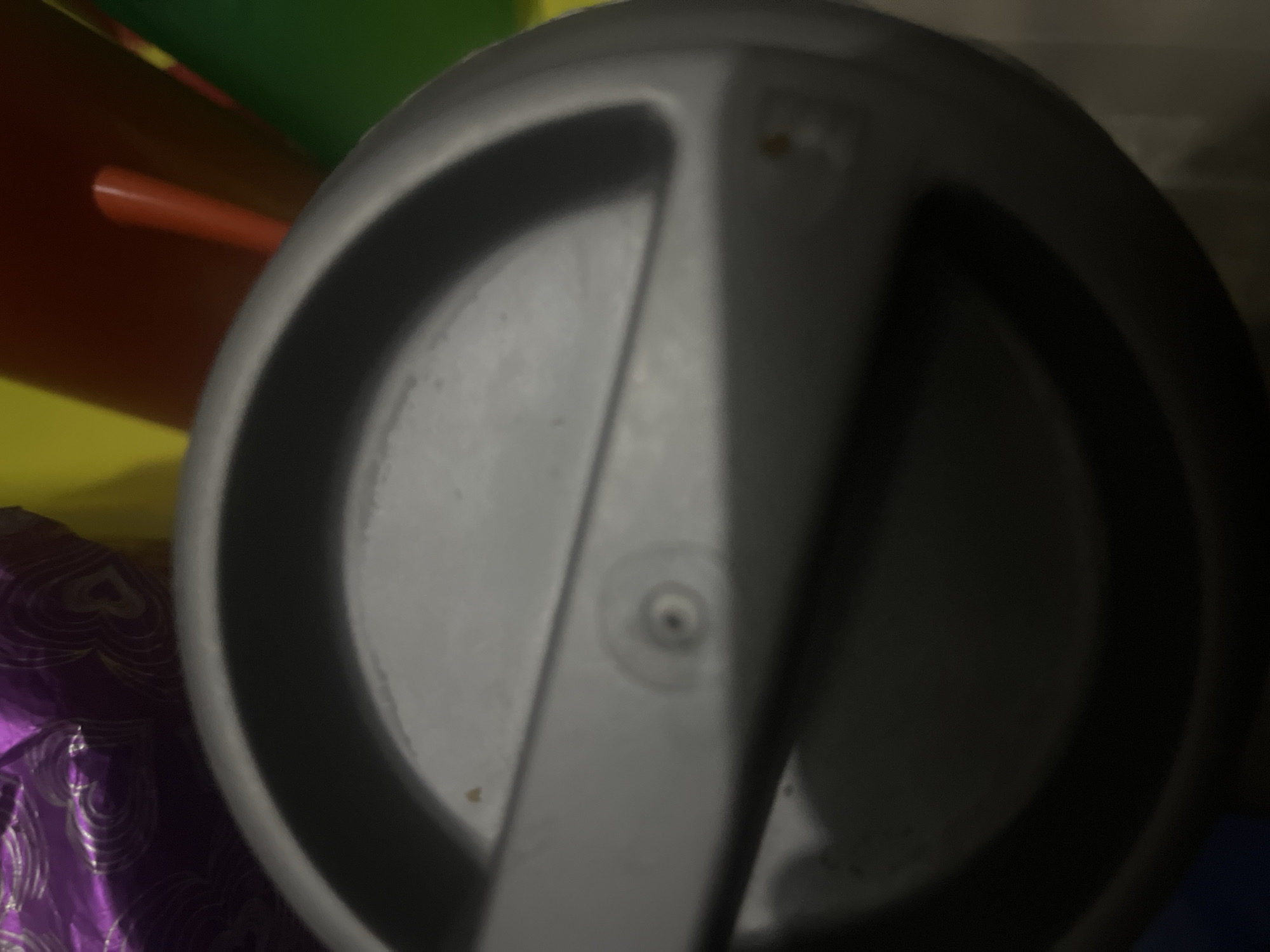 The image is a close-up, slightly blurry, overhead shot of a grey, lidded container, likely a thermal mug or a similar vessel. The lid has a slightly concave surface with a central raised circular detail. The container appears to be made of plastic. In the background, out of focus, are colorful objects, including a shimmering purple material with a swirling pattern, a bright yellow object, a red cylindrical object, and a green object. The lighting suggests an indoor environment. There are no discernible geographical landmarks to indicate a specific location within Jalingo, Nigeria.
