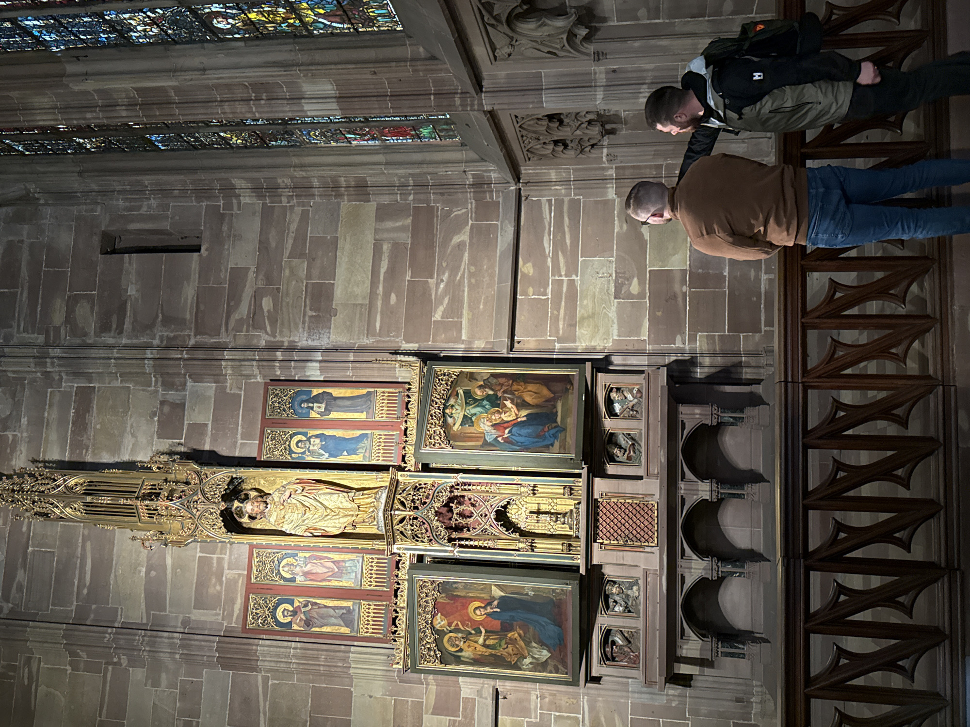 The scene depicts the interior of a Gothic cathedral, specifically the Cathédrale Notre-Dame de Strasbourg, France. The central focus is a large, ornate altarpiece positioned against a wall constructed of reddish-brown sandstone blocks.
The altarpiece features a tall, gilded, spired canopy at its apex. Below this, a large golden statue of a standing male figure, presumably Christ, is housed within a gilded, architectural niche. Flanking this central statue are four vertically oriented painted panels, two on each side, depicting individual religious figures or saints. Below the upper tier, the altarpiece expands horizontally. A central, smaller gilded Gothic structure is flanked by two large rectangular painted panels. The left panel shows an angel addressing a kneeling female figure; the right panel depicts multiple figures, including a woman with a child, in an indoor setting. The lower section of the altarpiece comprises a carved wooden or stone base with a central grilled compartment and several smaller carved busts or figures in niches. Below these, three empty arched niches are visible.
To the right of the altarpiece, a large Gothic stained-glass window is visible, composed of numerous colorful glass panes arranged in intricate patterns. A narrow, vertical slit-like opening is present in the stone wall to the upper left of the altarpiece.
In the foreground, two adult males stand behind a dark wooden balustrade with geometric cutouts, observing the altarpiece. Both have their backs mostly to the viewer. The man on the left has short, light brown hair, wears glasses, a brown long-sleeved top, and blue jeans, with his hands clasped behind his back. The man on the right has dark hair and a beard, is wearing a dark green vest over a dark long-sleeved shirt, dark trousers, and a dark backpack on his left shoulder. He is extending his right arm with his index finger pointed towards a detail on the altarpiece, appearing to indicate something to the other man.