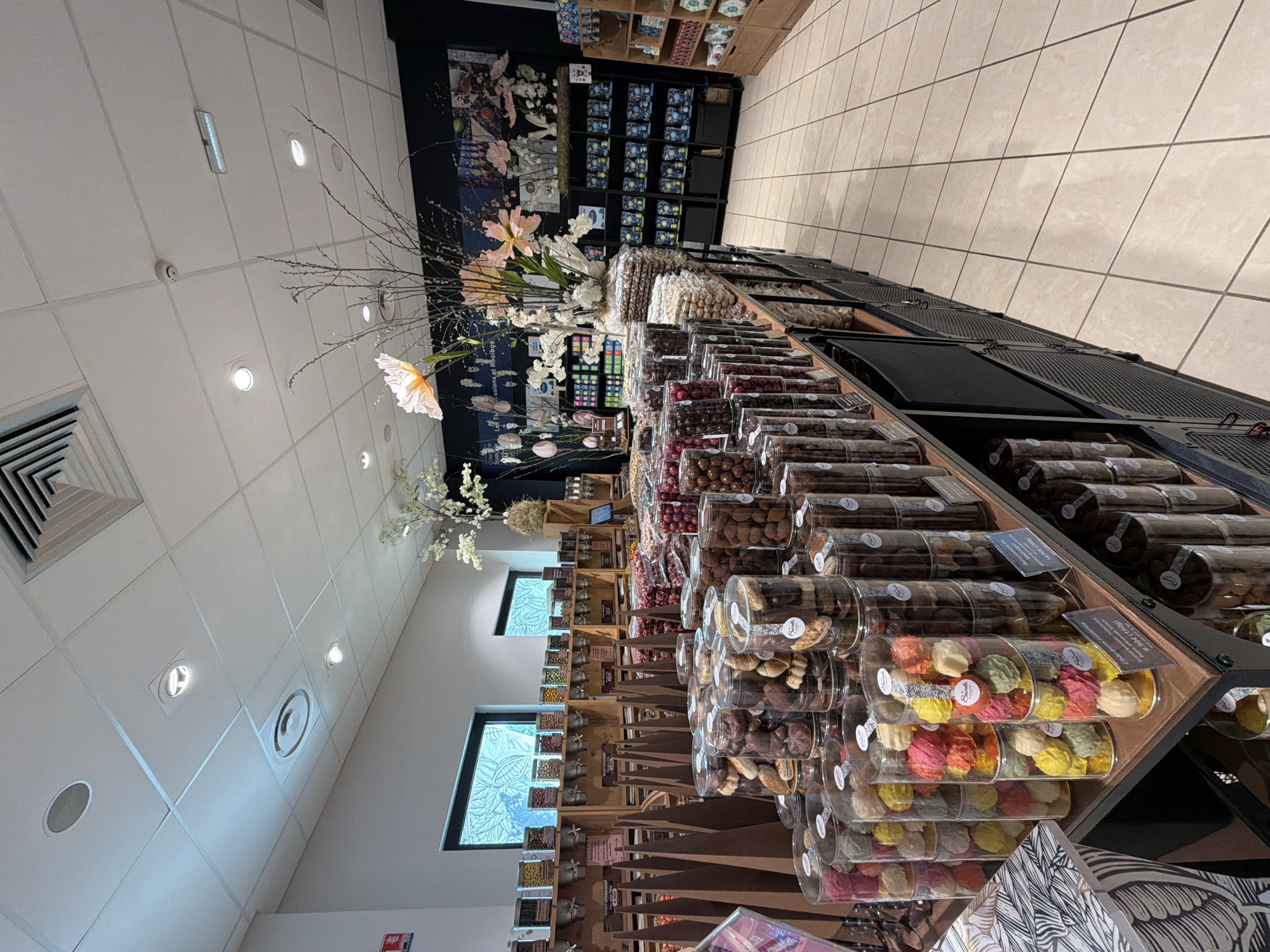 This image captures the interior of a brightly lit confectionery shop in Illkirch-Graffenstaden, France, presented from a perspective looking down a long display counter. The main subjects are the extensive array of packaged sweets, chocolates, and cookies, meticulously arranged in clear cylindrical containers. These containers are stacked on multi-tiered, dark-colored display units that extend into the background. Products include colorful meringue-like cookies, chocolate-covered nuts, and various other confectionery items, many bearing labels with a partial brand name such as "Schuler's." Small price tags or informational labels, like "3,90 €," are visible on some shelves.
The shop's setting combines modern retail design with artistic elements. To the left (in the corrected orientation), a dark accent wall features decorative floral arrangements with thin branches, white artificial flowers, a large paper butterfly, and smaller pink floral elements, alongside what appears to be a mural or large print depicting nature-inspired imagery. Below this, some boxed items are displayed on shelves, with partially visible text "Les illusions et l'odelba..." on the wall. Further back on this side, two rectangular windows with decorative, possibly etched or stained-glass patterns, provide ambient light.
The ceiling is composed of white grid tiles, fitted with numerous recessed circular lights, a rectangular emergency light, and a smoke detector, ensuring ample illumination. HVAC vents, including a large rectangular one and a circular one, are also visible. To the right, a light-colored tiled wall with a grid pattern extends into the distance, complementing the similar light-toned tiled floor. The overall impression is that of a clean, well-organized, and inviting specialty food store, likely open during the daytime given the bright indoor lighting. No people are visible, suggesting a quiet moment within the establishment.