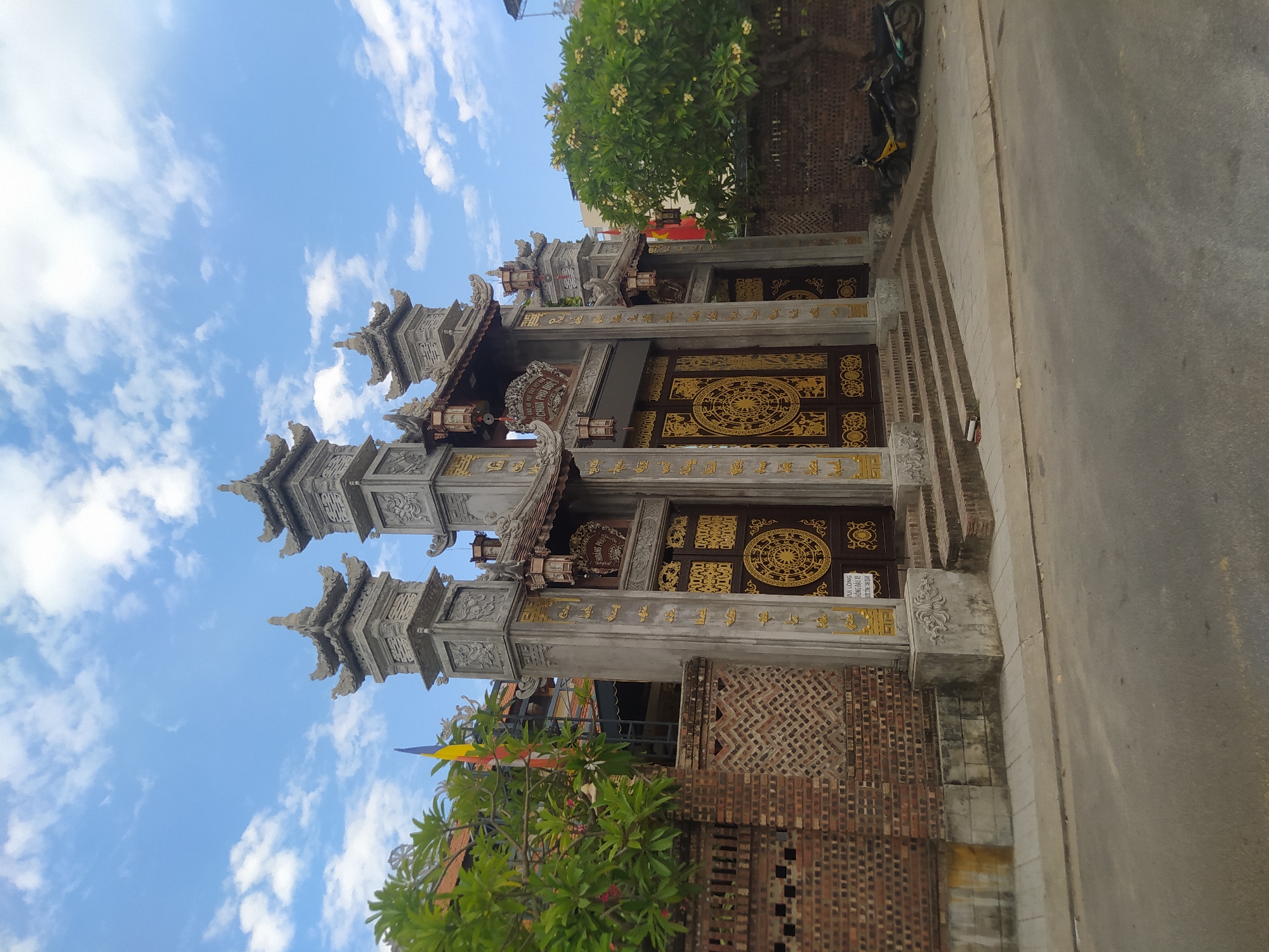 The image captures a segment of a temple entrance in Nha Trang, Vietnam, under a bright, cloudy sky. The architecture is ornate, with two imposing grey stone pillars framing a large, richly decorated wooden door. The pillars are topped with pagoda-style roofs, and both pillars and doors are adorned with intricate gold patterns and text. Small lanterns hang from the roofline above the entrance.
To the left of the entrance, a vibrant green tree with small white flowers provides a splash of nature against the stone and brick. Colorful prayer flags, a common sight at Vietnamese temples, are visible, adding to the cultural context. The entrance itself is set back from a paved road, which is visible on the right side of the image. A section of a brick wall, part of the temple's perimeter or adjacent building, is also visible.
On the road, a parked motorcycle is partially visible, suggesting the presence of visitors or residents. The overall scene evokes a sense of spiritual tranquility and cultural heritage. The bright daylight and scattered clouds indicate it is daytime, likely mid-morning or afternoon. No people are directly visible in the frame.
Visible text on the door and pillars appears to be in Vietnamese. One sign clearly reads "CHÙA PHƯỚC LỘC" which translates to "Phuoc Loc Pagoda". Another sign near the entrance has text that is partially obscured but appears to contain Vietnamese characters and possibly numbers. The ornate designs suggest a place of worship that is well-maintained and visually striking.