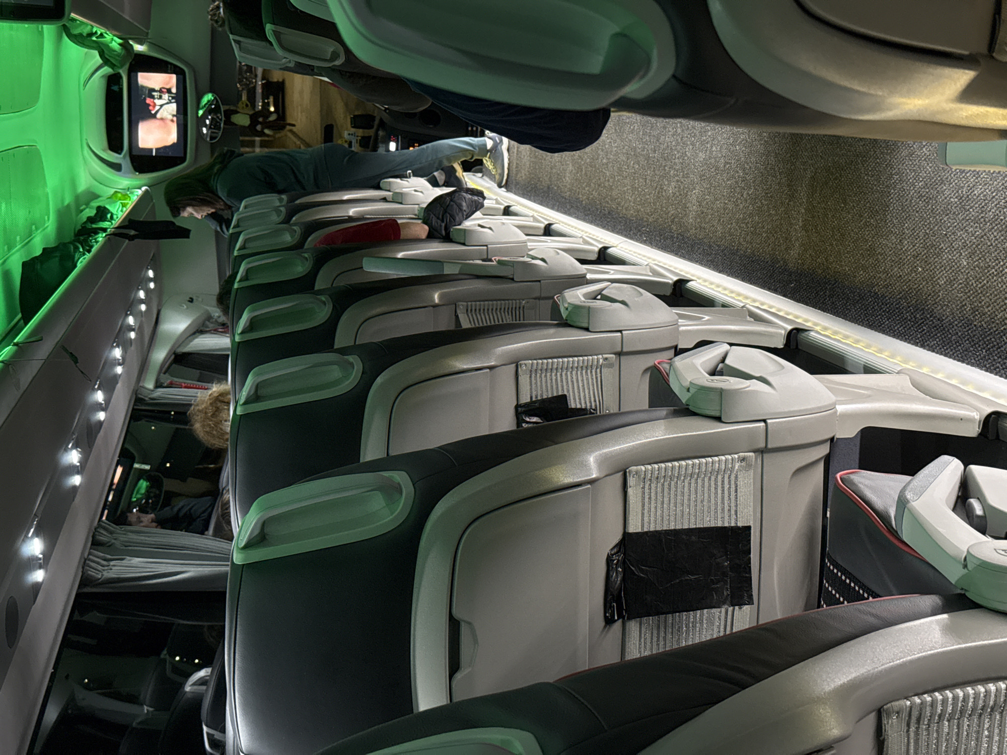 The image shows the interior of a sleeping car on a train. The car has multiple rows of bunk beds, with several people occupying them. A person wearing a green shirt is lying on a bed watching a television screen mounted on the wall. Some of the bunks appear to be occupied by sleeping passengers. The interior is illuminated with green and white lighting. The car’s seats are modern, with grey and black accents and built-in headrests. The location is unknown and there is no indication of being in the city of Veliko Tarnovo, Bulgaria.