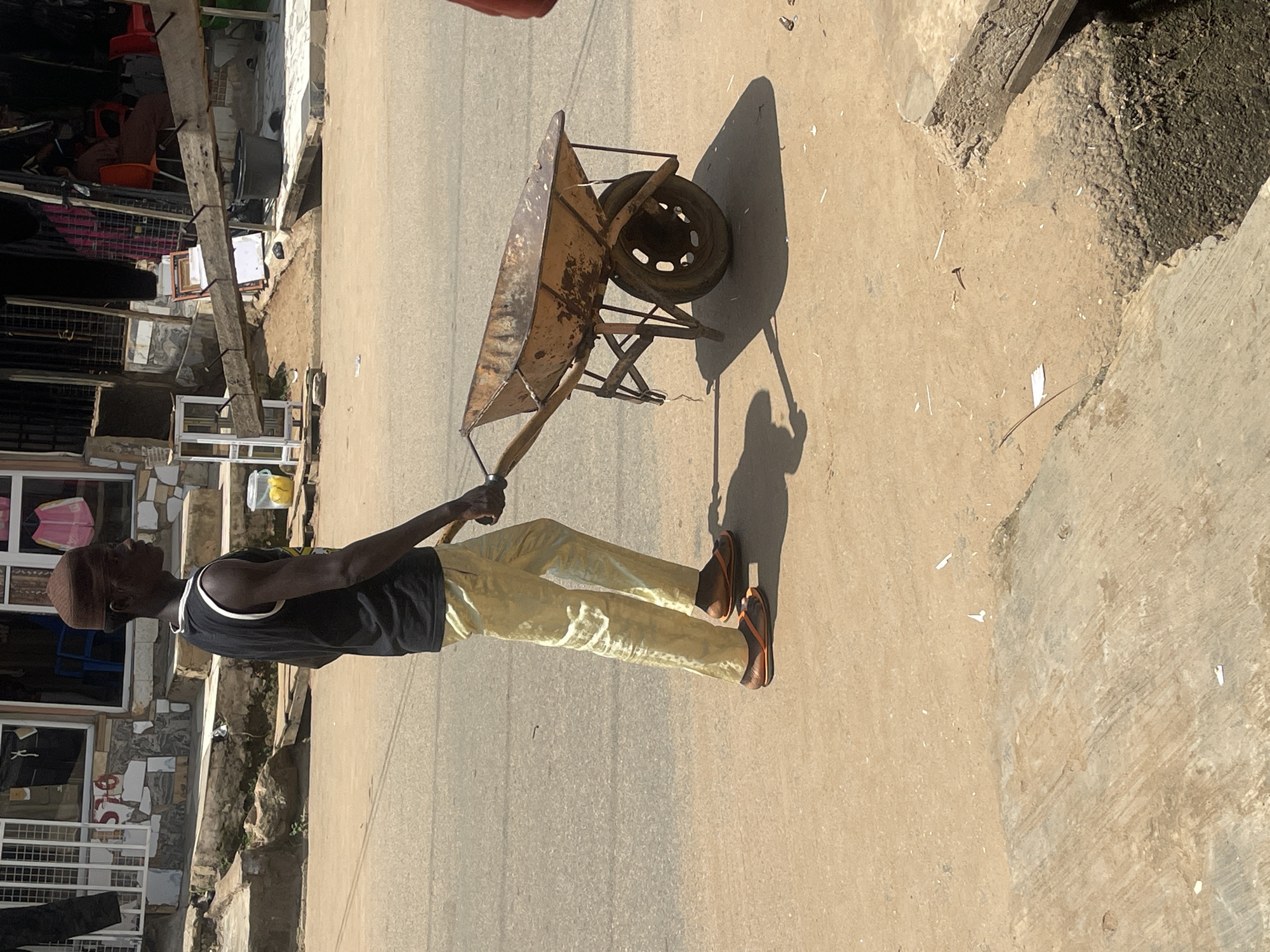 A man wearing a dark sleeveless shirt and light yellow trousers walks from left to right, pushing a rusty, two-wheeled wheelbarrow on a paved road. He is also wearing an orange flip-flop on his right foot and a similar one on his left. He is in profile and looking to the right. The wheelbarrow appears to be empty. Behind the man, a building with a tiled facade, white metal window grills, and an open doorway is visible. The area appears to be a street in Abuja, Nigeria, with a dusty, sunlit environment. In front of the building, there are steps and some debris on the ground.