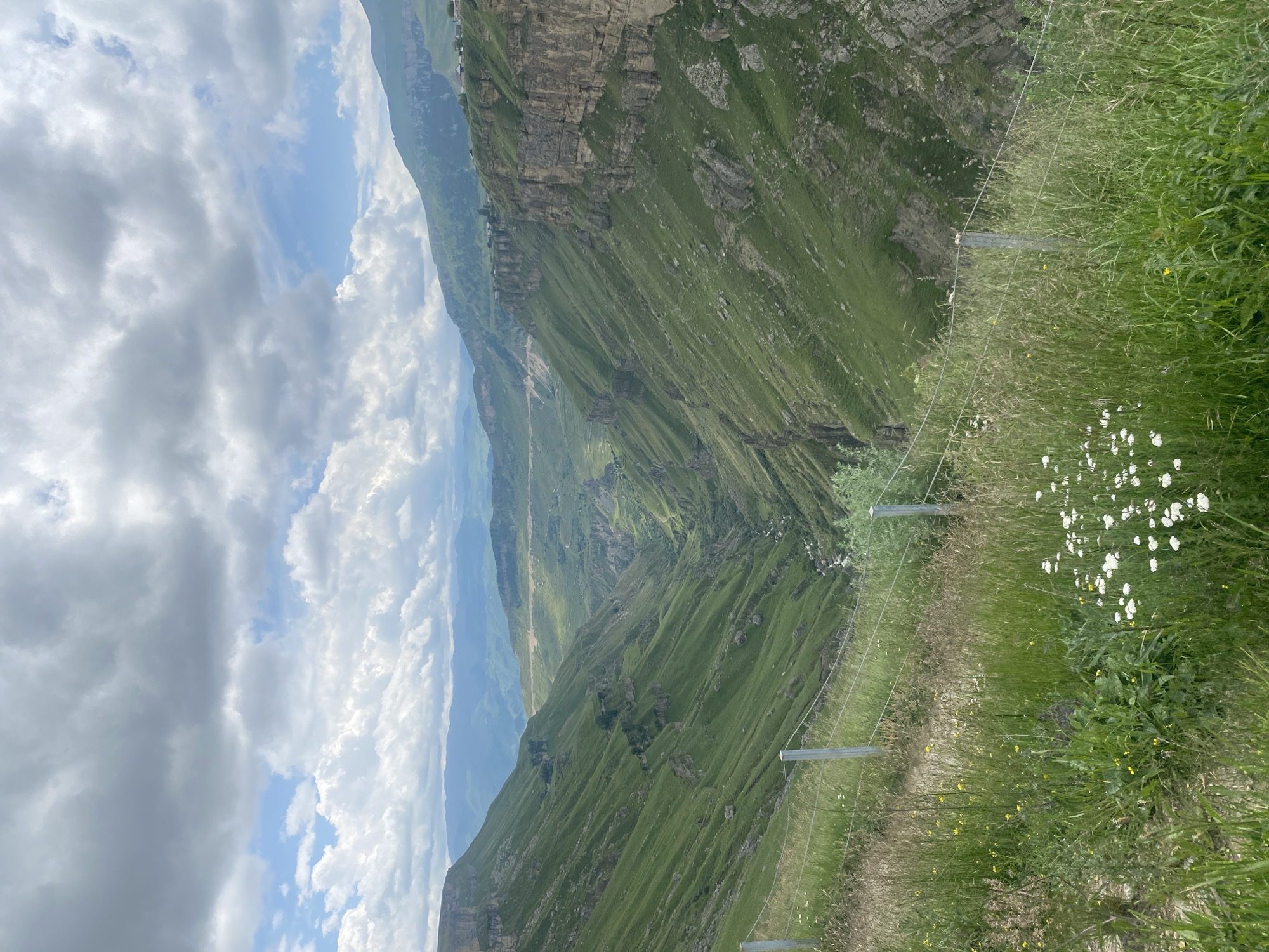 The image shows a panoramic view of a deep, verdant gorge with steep, grassy slopes. In the foreground, a fence made of metal posts and wires runs horizontally across the frame, separating a patch of wildflowers and tall grass from the precipice. The gorge itself is characterized by dramatic cliffs and winding, green hills that descend into a narrow valley where a river or stream is visible. Distant mountains and a hazy sky with scattered clouds complete the landscape. The scene is devoid of any people or animals. The location is identified as the city of Khunzakh, Russia, known for its dramatic natural scenery.