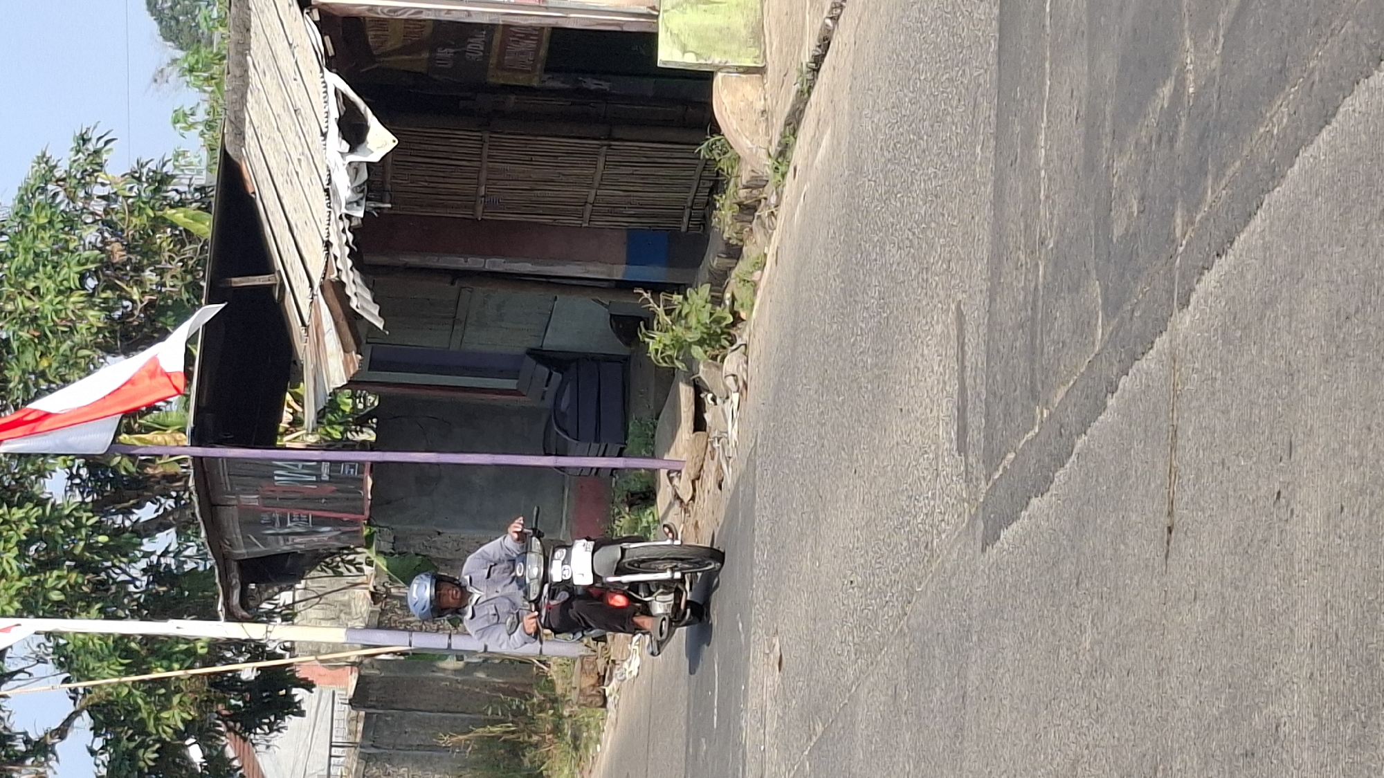 A person wearing a grey shirt and a blue helmet rides a white motorcycle on a paved road. The person is on the left side of the road, facing forward. The motorcycle has a white front panel and black tires. Behind the motorcycle rider, there is a small building with a corrugated metal roof. A red, white, and blue Indonesian flag is visible on a pole in front of the building. The environment appears to be a roadside in a rural or suburban area, possibly in Cilaku, Indonesia, with some vegetation and debris scattered along the side of the road. The sky is clear and blue.