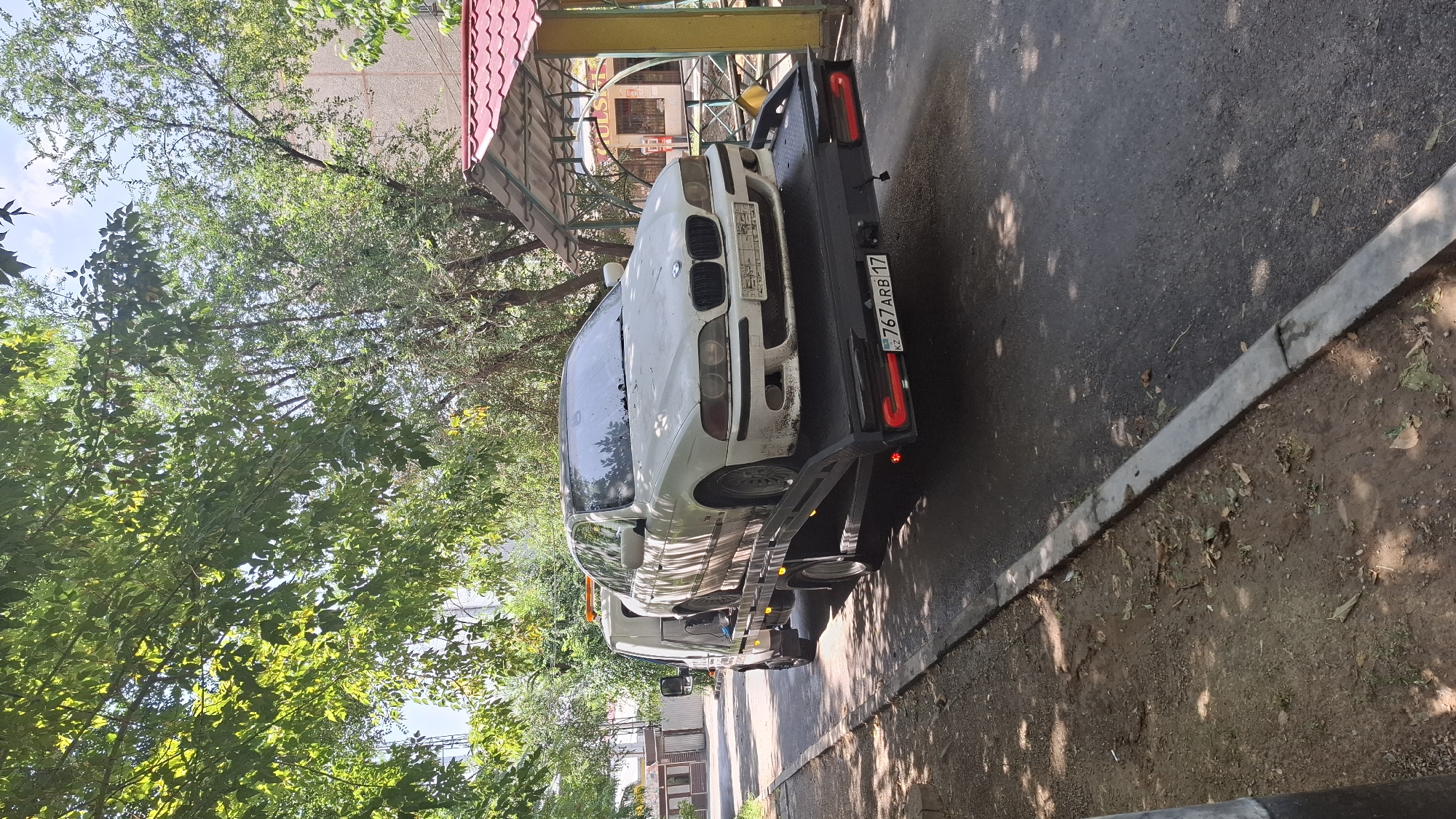A white BMW sedan is loaded onto a black tow truck on a paved road in Shymkent, Kazakhstan. The BMW is facing forward on the tow truck's flatbed, with its front end slightly elevated. The tow truck's hazard lights are illuminated. Trees with green foliage line the left side of the road, casting shadows on the ground. In the background, a building with a red-tiled roof structure is visible. The license plate of the BMW reads "KZ 767 ARB 17". The scene appears to be a street in a residential or commercial area.