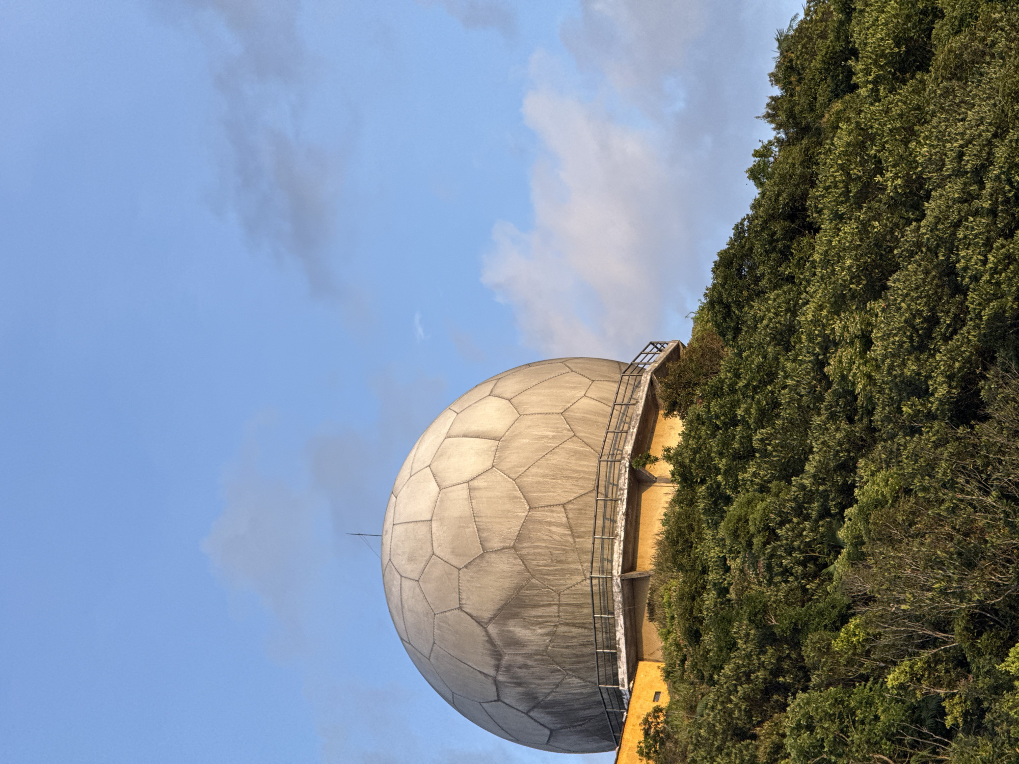 The image shows a large, grey, geodesic dome structure situated on a lush, green, tree-covered hillside. The dome is segmented into many polygonal panels and is supported by a concrete platform with a metal railing. A yellow building or structure is visible beneath the dome. The sky above is a clear blue with a few wispy clouds. This structure is the Đà Nẵng Meteorological Radar Station, located on Son Tra Peninsula in Đà Nẵng, Vietnam. There are no people or animals visible in the image. The scene depicts a clear day with the sun casting shadows on the hillside.
