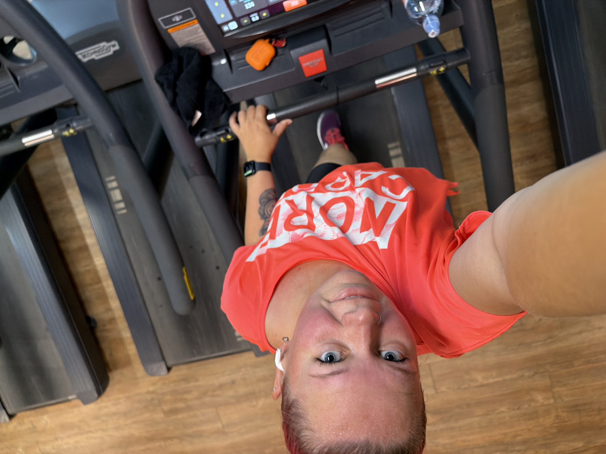 A woman with fair skin, light blue eyes, and pinkish-blonde hair is captured in a selfie taken from a high angle, looking up at the camera. She is wearing a bright coral-colored t-shirt with white lettering that reads "MOR" and has a nose ring and white wireless earbuds in her ears. She appears to be sweating, indicating she is likely engaged in physical activity. She is holding onto the handle of a treadmill, suggesting she is at a gym. The treadmill is black with silver accents and a digital display console. An orange case, possibly for earbuds, and a red object are on the console. A black cloth and a water bottle are also visible near the console. The floor beneath the treadmill is made of light brown wood planks. The image is oriented vertically, with the woman's head towards the top left corner. The overall scene depicts a workout session at a gym.
