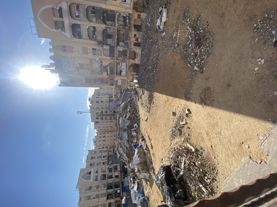 The image depicts a severely damaged urban landscape in Khan Yunis, Palestinian Territory, under a clear blue sky with bright sunlight emanating from the upper right, casting distinct shadows. Multiple multi-story residential buildings, constructed of light-colored material, frame the scene. These buildings exhibit extensive structural damage, including large impact holes, shattered windows, exposed interiors, and darkened sections consistent with explosions or fires. Cellular communication antennas are visible on the rooftops of several structures.
The ground level is largely barren earth and sand, covered with significant debris. A vast mound of rubble, concrete fragments, and twisted metal occupies the central background, indicating the complete collapse of one or more structures. To the left mid-ground, a cluster of makeshift shelters, constructed from tarpaulins and various materials, are erected on the dusty ground amid scattered refuse. In the right mid-ground, substantial piles of debris include the recognizable, heavily damaged chassis of a white vehicle. The immediate foreground features a section of reddish interlocking pavers in the bottom left corner, alongside bare earth and minor debris. No people are visible, and no active interactions or actions are depicted.