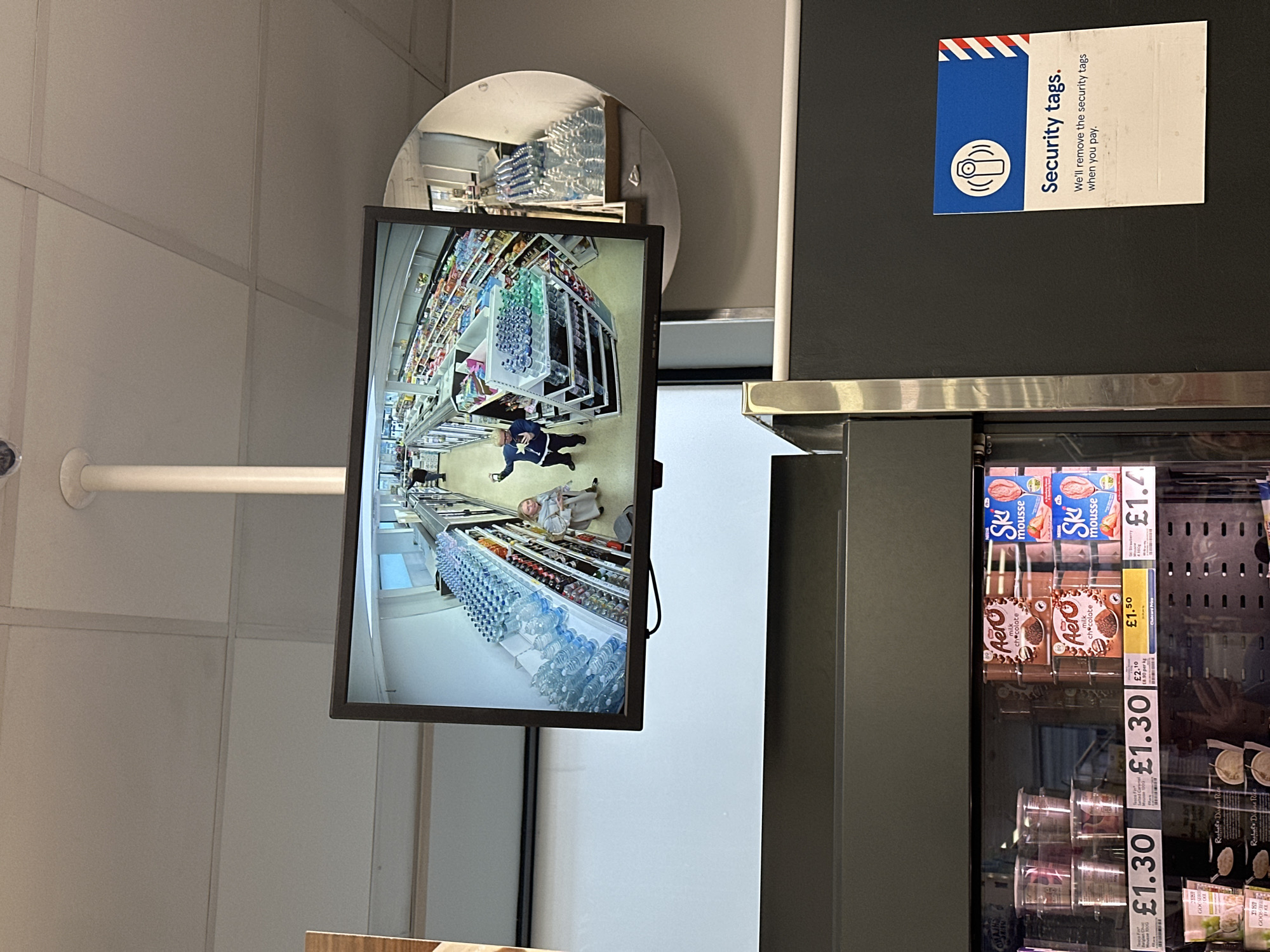 This is an indoor shot of a retail environment. A security camera display screen is mounted on a wall, showing a live feed of the store aisle. Two individuals are visible in the camera's feed. The screen is mounted next to a refrigerated display case containing yogurt, Aero chocolate bars, and mousse. A sign stating "Security tags. We'll remove the security tags when you pay" is on the wall. The overall environment is brightly lit and appears to be in a store located in London, United Kingdom. The image does not provide any details of any objects interacting with the environment.