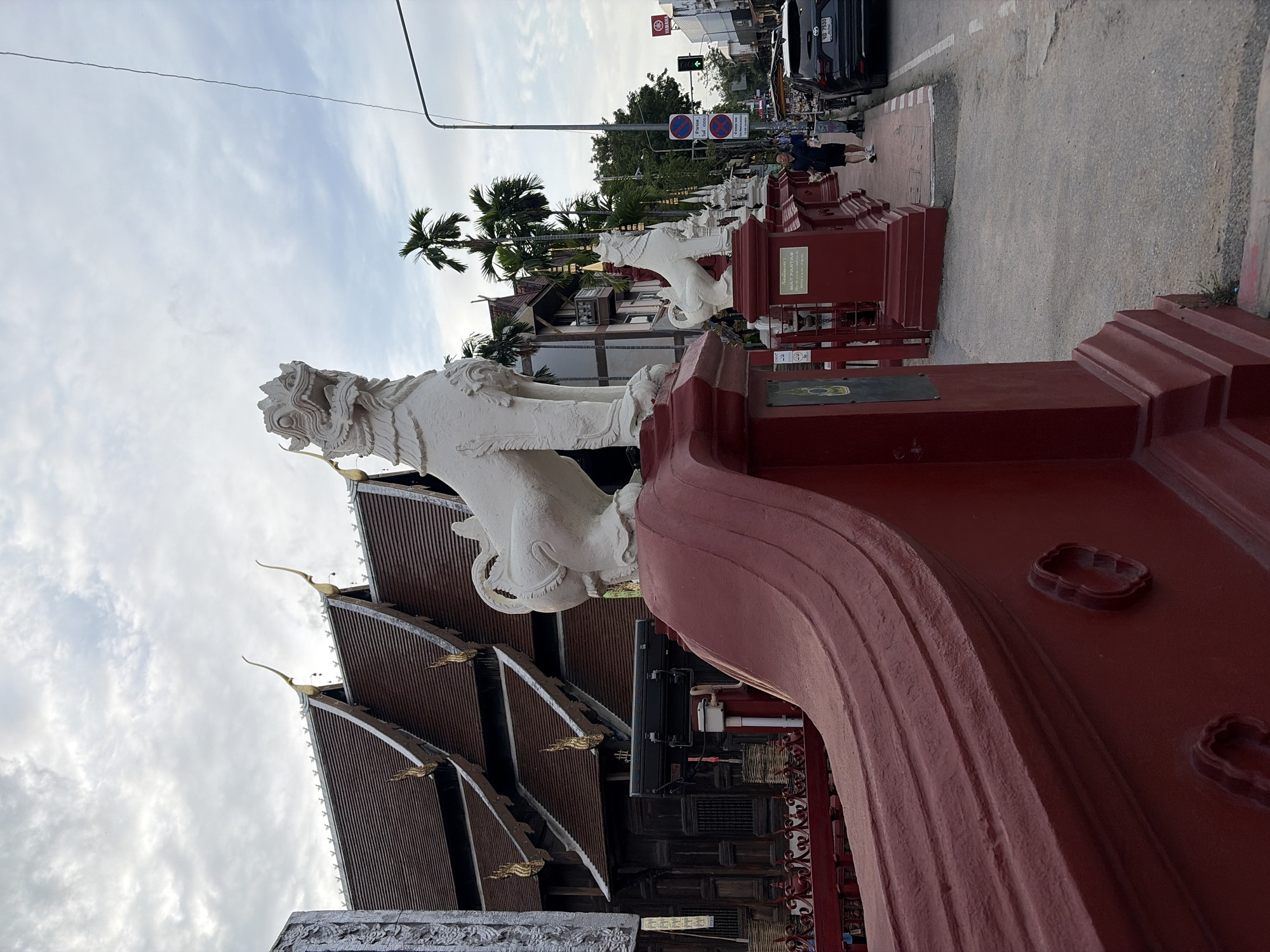 The image shows the entrance to a temple in Chiang Mai, Thailand, with traditional Thai architecture. In the foreground, there is a red ornate railing and a white lion statue. In the background, a wooden temple building with a multi-tiered roof and golden spires is visible. A street with cars and pedestrians runs alongside the temple complex. The sky is cloudy, suggesting overcast weather. The overall scene depicts a vibrant cultural and religious site within an urban setting.