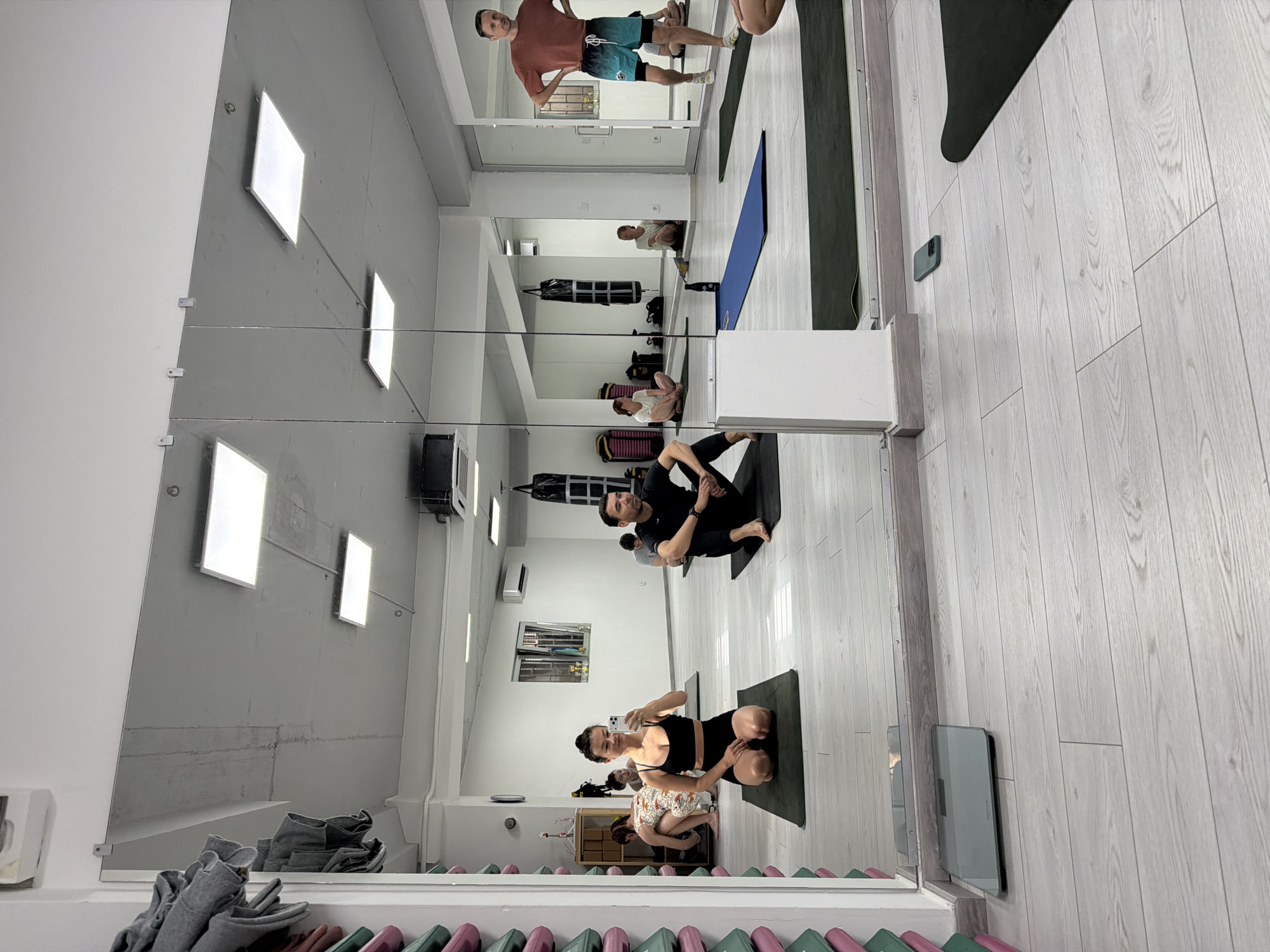 The image shows a yoga class in progress, reflected in a large mirror spanning the wall of a fitness studio. There are multiple participants, including one woman in the foreground wearing a black sports bra and shorts, holding a smartphone, likely taking a selfie. A man further back is practicing a seated yoga pose. Other participants are scattered throughout the room, engaged in different poses.
The studio has a white ceiling, gray walls with inset rectangular light panels, and light-colored wooden flooring. Yoga mats are spread on the floor. There are lockers and storage units along the wall. In the reflection, a doorway leads to another part of the fitness facility.
Based on the location of the image, the yoga class is most likely taking place in a fitness studio in the city of Budva, Montenegro.