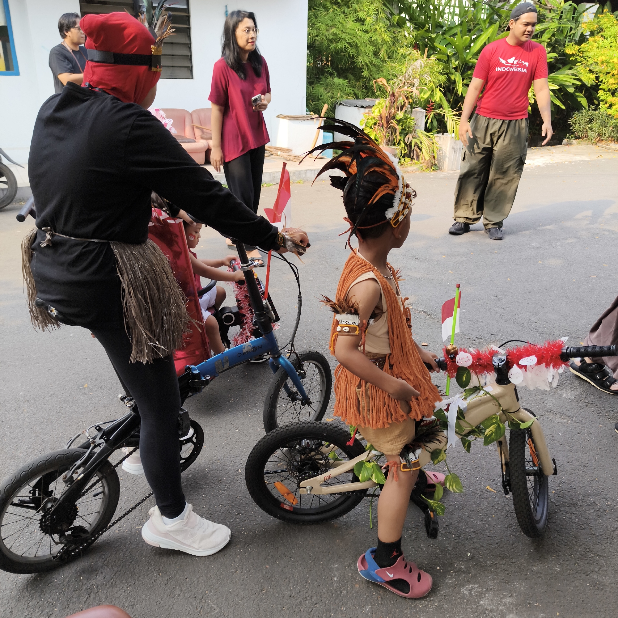 In a daytime outdoor scene on a paved surface, likely a street or parking lot, in South Jakarta, Indonesia, two children are positioned next to bicycles. The child on the left, wearing a black outfit with a red head covering and a fringed skirt, is seated on a blue bicycle. This child also wears black leggings and white sneakers. The child on the right is standing and wearing a costume that includes an orange fringed tunic, armbands adorned with feathers and shells, and a headdress with feathers and shells. This child is standing next to a cream-colored bicycle decorated with red garland, white ribbons, and greenery. A small Indonesian flag is attached to the handlebars of this bicycle.
In the background, several adults are visible. To the left, a man wearing a black t-shirt and dark pants stands with his back to the camera. Next to him, a woman in a maroon t-shirt and black pants, wearing glasses, holds a phone. To the right, a man in a red t-shirt with the word "INDONESIA" printed on it and olive green cargo pants, wearing a grey baseball cap, is looking to the side. The environment includes a white building with a window to the left and lush green foliage and trees in the background to the right. The overall scene suggests a cultural event or parade, possibly related to Indonesian Independence Day given the flag and "INDONESIA" shirt.