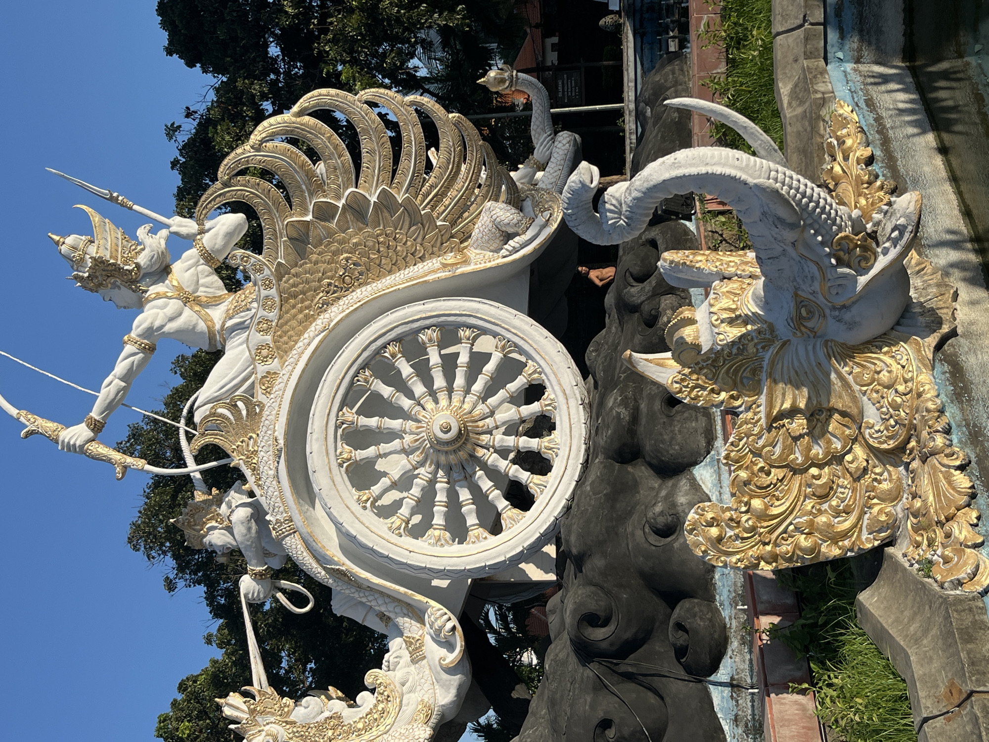 The image shows a large, ornate statue of a chariot with figures and mythical creatures. The statue is predominantly white and gold and appears to be made of concrete or a similar material. In the chariot, a male figure is depicted drawing a bow, while another figure, likely a driver, is also present. The chariot itself has elaborate winged decorations and a large spoked wheel. The chariot rests on a dark, cloud-like base. In the foreground, there is a white elephant statue with gold accents, its trunk raised. The statue is situated in an outdoor environment with trees and a clear blue sky in the background. A person can be seen standing in the shadow of the chariot. The statue is located at the statue of the Mahabharata in Kuta, Indonesia.