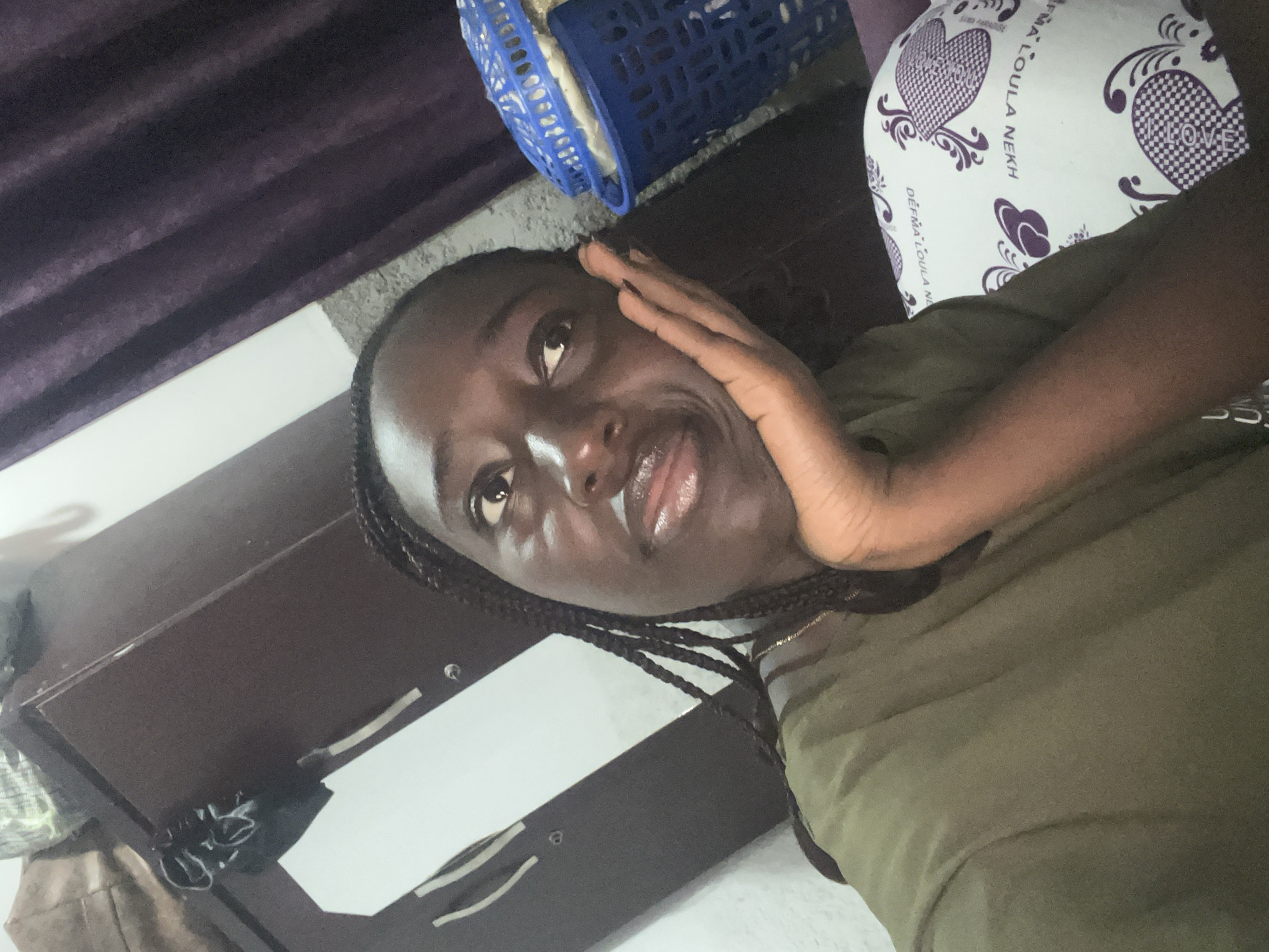 The image presents an indoor scene, likely a bedroom, in the city of Abuja, Nigeria. A young adult Black woman is depicted in a close-up, head-and-shoulders shot, angled slightly upwards. She has braided hair and is wearing a dark olive green short-sleeved top and a thin gold-colored chain necklace. Her chin rests on her right hand, held with an open palm, and her eyes are cast upwards and to the viewer's left, indicating a contemplative or pensive expression. Her skin exhibits a subtle sheen, particularly on her forehead, nose, and lips.
In the background to the woman's left, a dark brown wooden wardrobe is visible, featuring two light-colored vertical embellishments that may be handles. Several indistinct items rest on the top surface of the wardrobe. To the woman's right, dark purple curtains with vertical pleats extend upwards. Adjacent to her head and partially obscured is a bright blue plastic laundry basket with a matching lid, containing light-colored fabric items. In the lower right corner, a white pillow is partially visible, decorated with purple hearts (some solid, some checkered) and intricate scrollwork, along with the text "DERMA LOULA NEKH" and "LOVE."