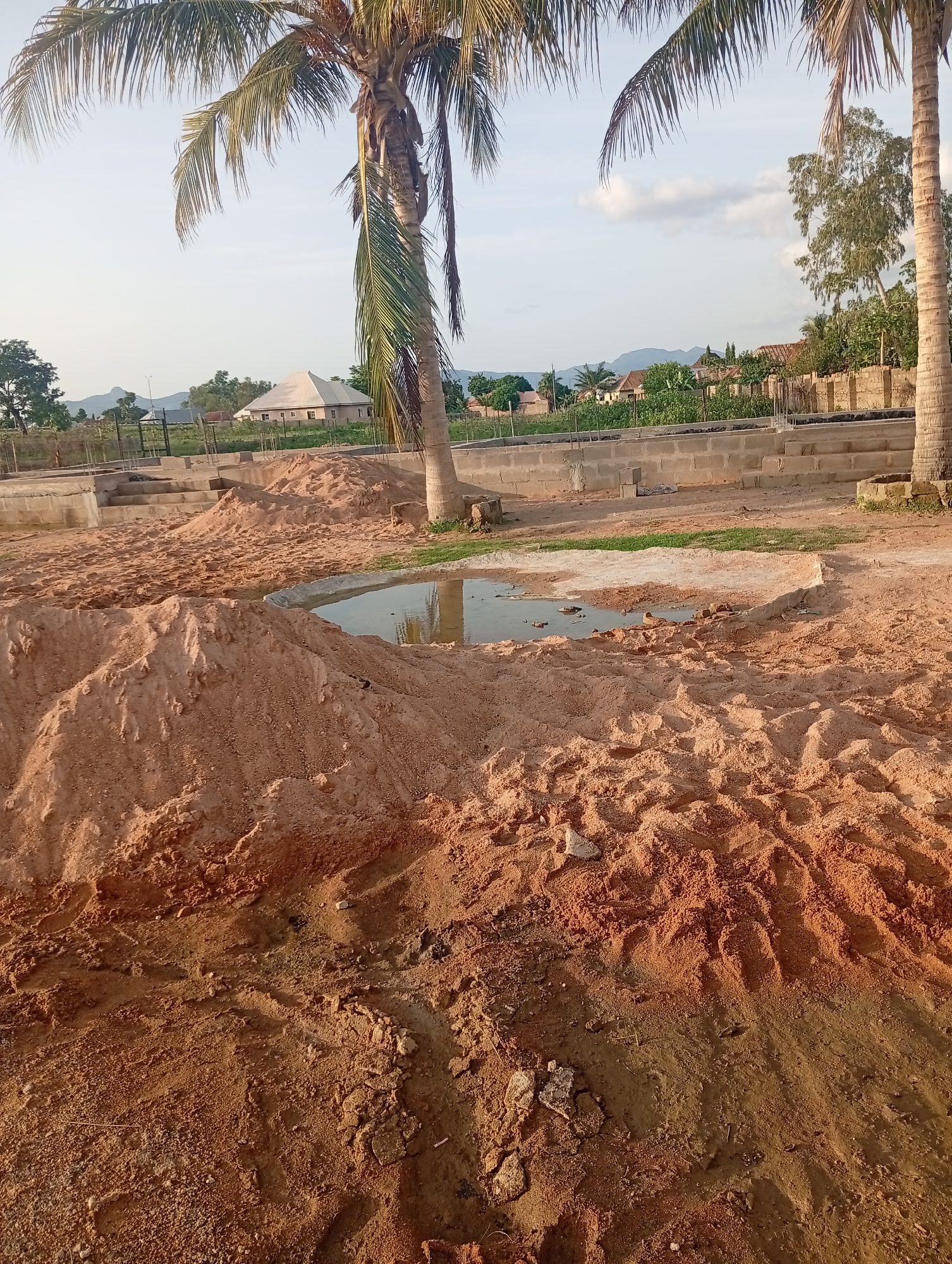 The image captures an outdoor scene at what appears to be a construction site in Jalingo, Nigeria. The foreground is dominated by piles of reddish-brown sand, some of which have been disturbed, suggesting recent activity. A shallow pool of water reflects the sky and surrounding elements, indicating recent rainfall or the presence of a water source.
In the middle ground, the foundation of a building is visible, constructed with concrete blocks. Reinforcing bars protrude from the structure, further indicating ongoing construction. Beyond the immediate construction area, several buildings with pitched roofs can be seen, some appearing residential. A low wall, likely a property boundary, is also present.
Two prominent palm trees frame the scene, with their fronds reaching towards the sky. Their presence, along with the general landscape, suggests a tropical or sub-tropical environment. In the distance, hills or mountains are visible under a hazy sky with scattered clouds. The lighting suggests it is either early morning or late afternoon, with the sun casting long shadows and bathing the scene in a warm glow.
The overall atmosphere is one of development and transition, with the raw materials of construction juxtaposed against existing structures and natural elements. There is no visible text in the image. The image does not contain any people.