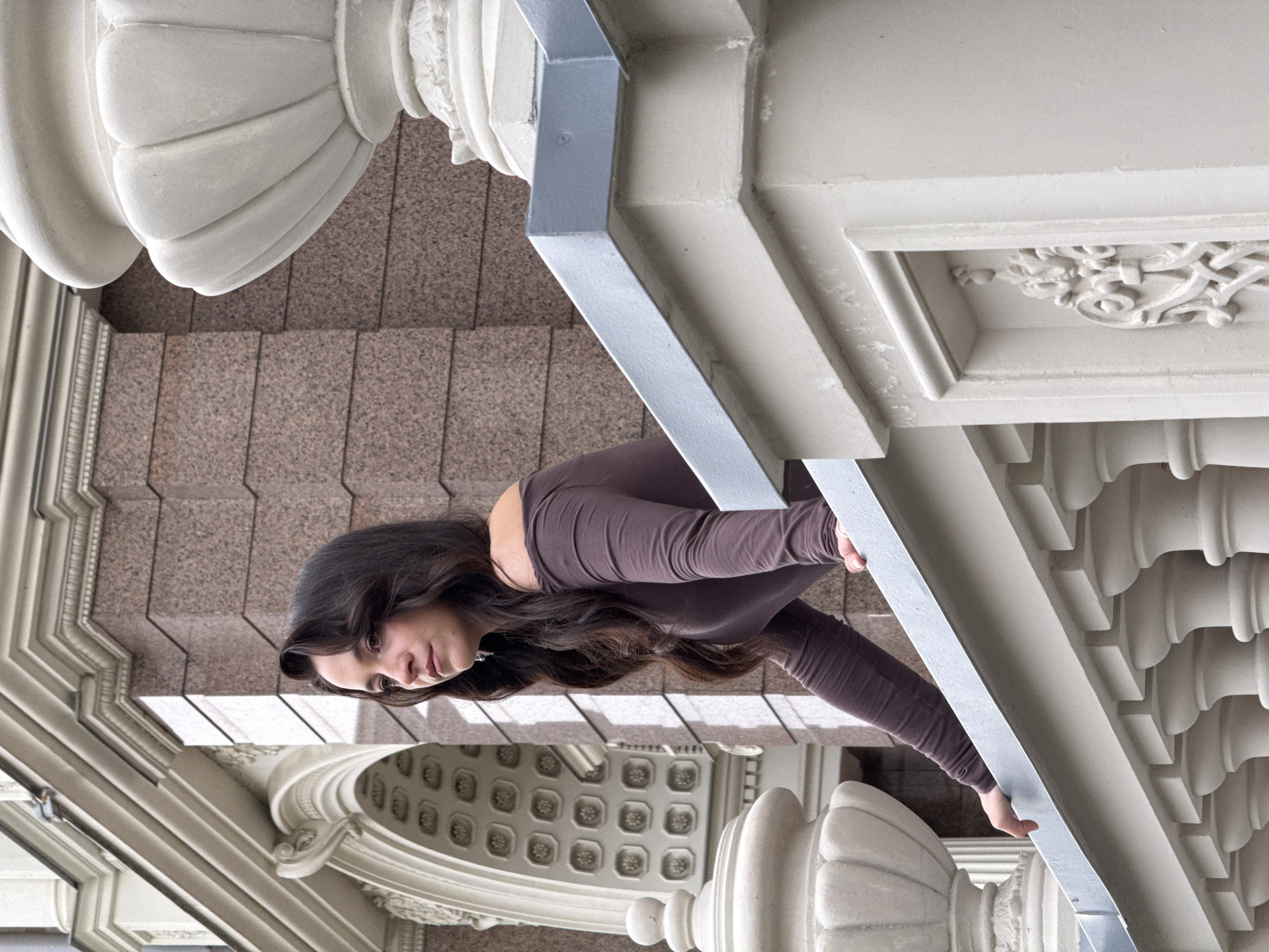 A young woman with long, dark, wavy hair leans over a light-colored stone balustrade. She is wearing a long-sleeved, off-the-shoulder brown top. The balustrade has decorative stone elements, including urns and ornate carvings. Behind her is a building with a textured, reddish-brown facade and detailed architectural features, including an arched entrance with decorative insets. The scene appears to be an outdoor architectural setting.