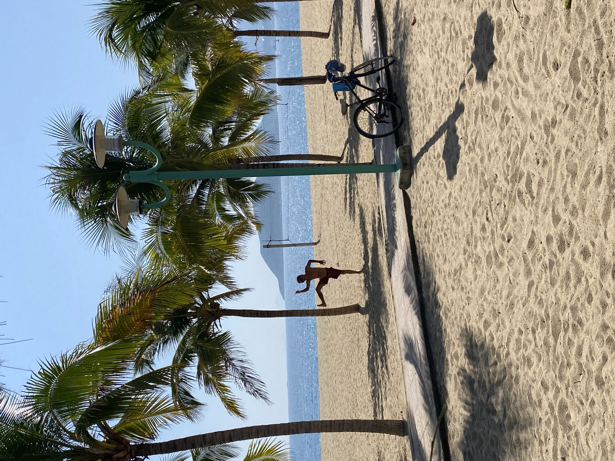 The image depicts a sunny day at a beach in Nha Trang, Vietnam. In the foreground, there is a sandy beach with palm tree shadows cast across it. A paved walkway runs along the edge of the sand. On the walkway, a bicycle is parked, leaning against a light post. The light post is green with two lamps. Further back, on the sand, a young man is captured mid-action, performing a kick, with one leg extended. Behind him is the ocean, a tranquil blue body of water, and a sandy beach that stretches into the distance. The horizon is marked by the meeting of the sea and the sky, with a faint outline of a distant landmass. A volleyball net is visible on the beach in the middle ground. The scene is characterized by the lush green of the palm trees and the bright blue of the sky and sea, suggesting a typical tropical beach environment.