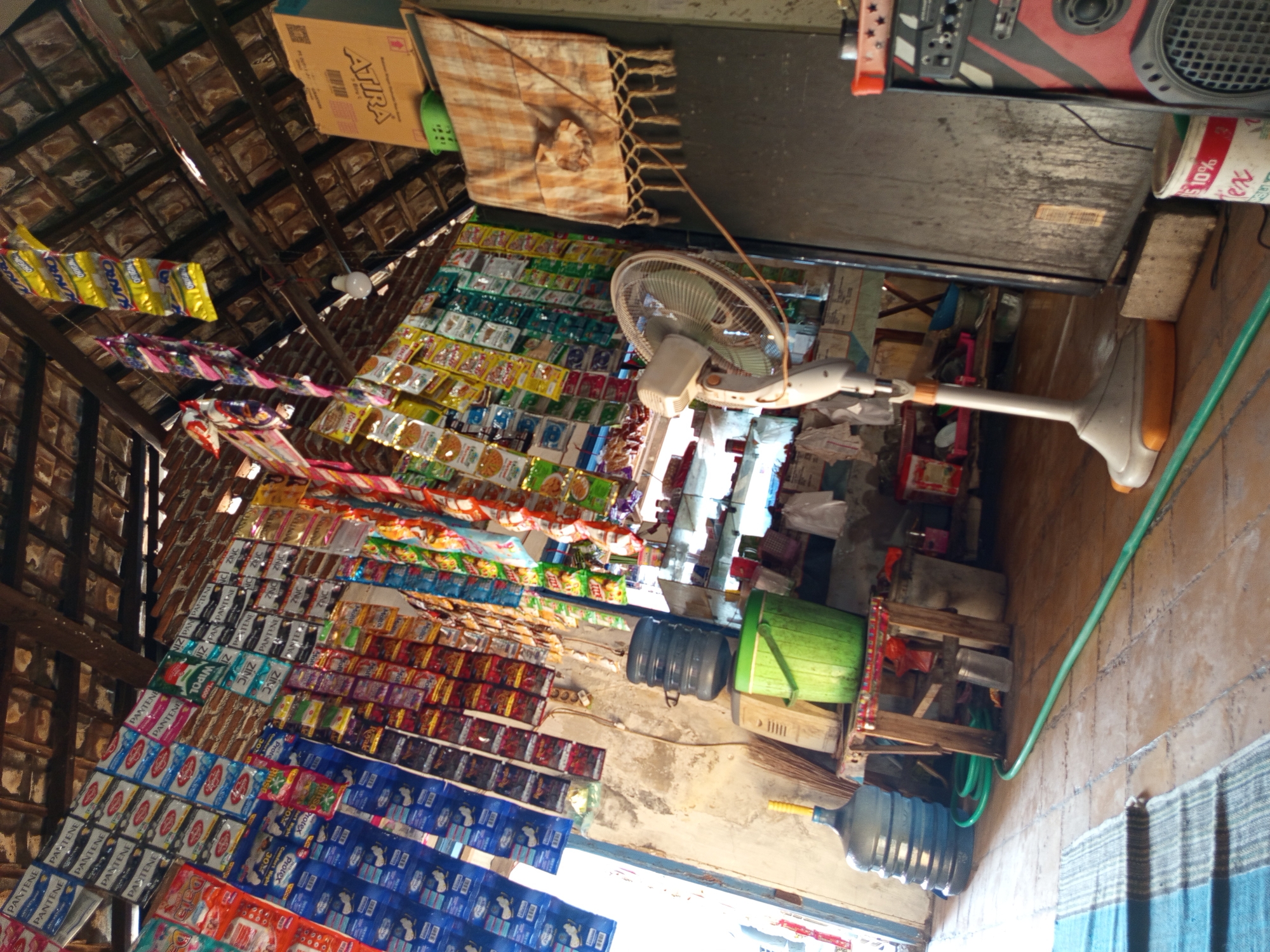 The image depicts the interior of a small, rustic shop in Tebel, Indonesia. The shop is packed with various merchandise, predominantly sachets of food and personal care products, displayed on strings hanging from the thatched roof and a brick wall. In the foreground, a large blue water jug sits on the floor next to a green garden hose. Further into the shop, a stack of smaller water containers is visible, as well as a green bucket and a broom leaning against the wall. A white standing fan is positioned in the center of the frame, and a dark refrigerator or cabinet stands to its right. A piece of patterned cloth with fringes hangs on the wall behind the fan. A cardboard box is placed on top of the cabinet. In the background, shelves are visible, stocked with more items. The floor is made of terracotta tiles. The lighting suggests it is daytime, with natural light entering from an open doorway or window on the left. There are no people present in the image.