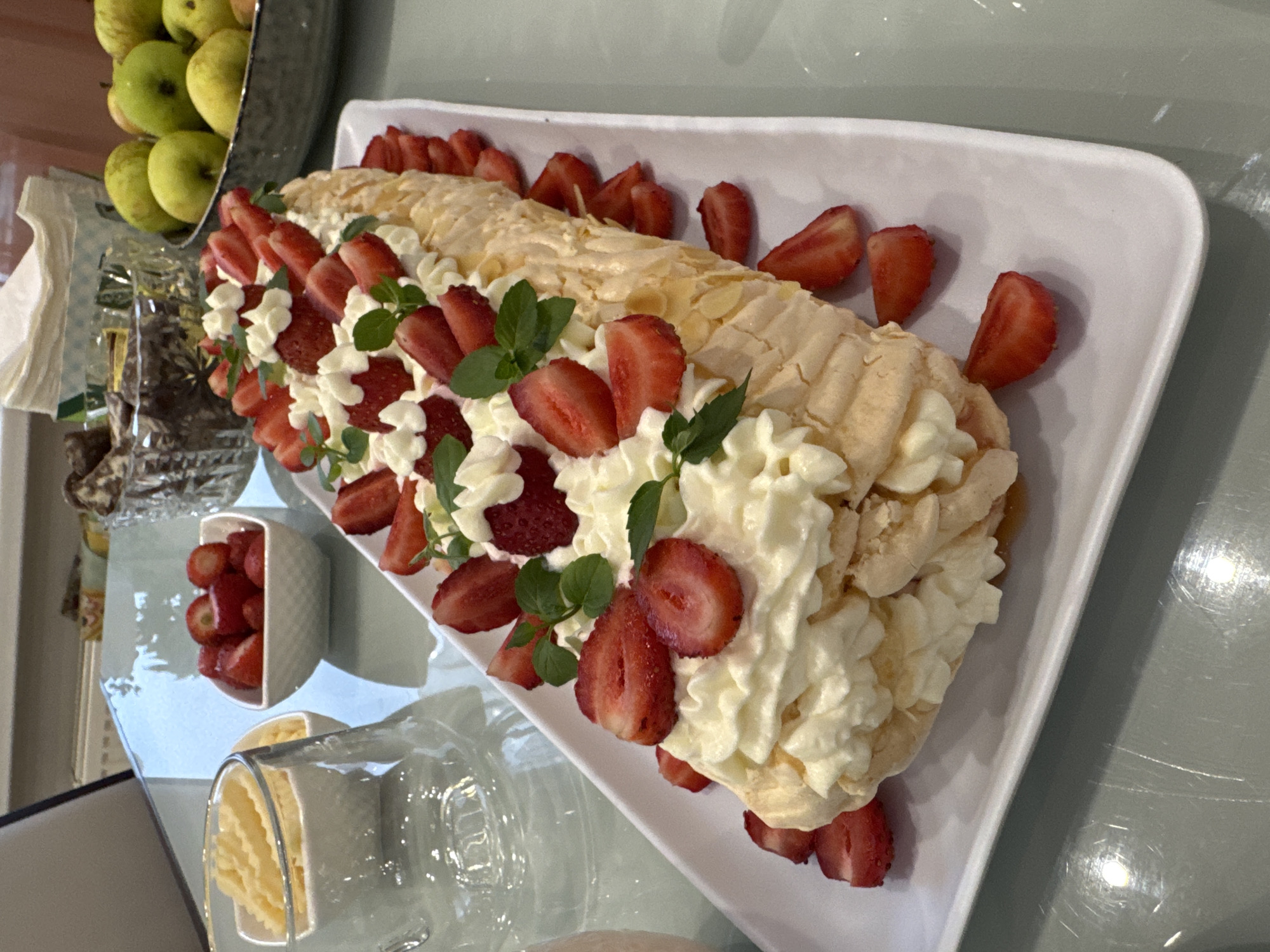 The image shows a festive dessert, likely a Pavlova roll, elegantly displayed on a white rectangular platter. The dessert is generously topped with whipped cream and fresh strawberries, with a few mint leaves adding a touch of green. The base of the dessert appears to be a crisp meringue, lightly golden brown, and possibly sprinkled with flaked almonds. Surrounding the main dessert are more sliced strawberries, creating a visually appealing presentation.
In the background, on the same glass-topped table, are several other items. To the left, a clear glass or pitcher is partially visible, and next to it, two small white rectangular bowls contain what appears to be crinkle-cut fries or a similar snack. Further to the right, a clear glass bowl is filled with dark chocolates or cookies. Beyond that, a rustic-looking bowl holds several green pears. A light-colored fabric napkin dispenser is also visible in the background. The overall setting suggests a casual gathering or celebration, possibly a meal or dessert course being served. The lighting indicates an indoor environment, with natural light possibly supplemented by artificial light, creating reflections on the glass tabletop.
The image does not provide any specific information about the city of Saint Petersburg, Russia, or any individuals present. The description is solely based on the visual elements within the photograph.