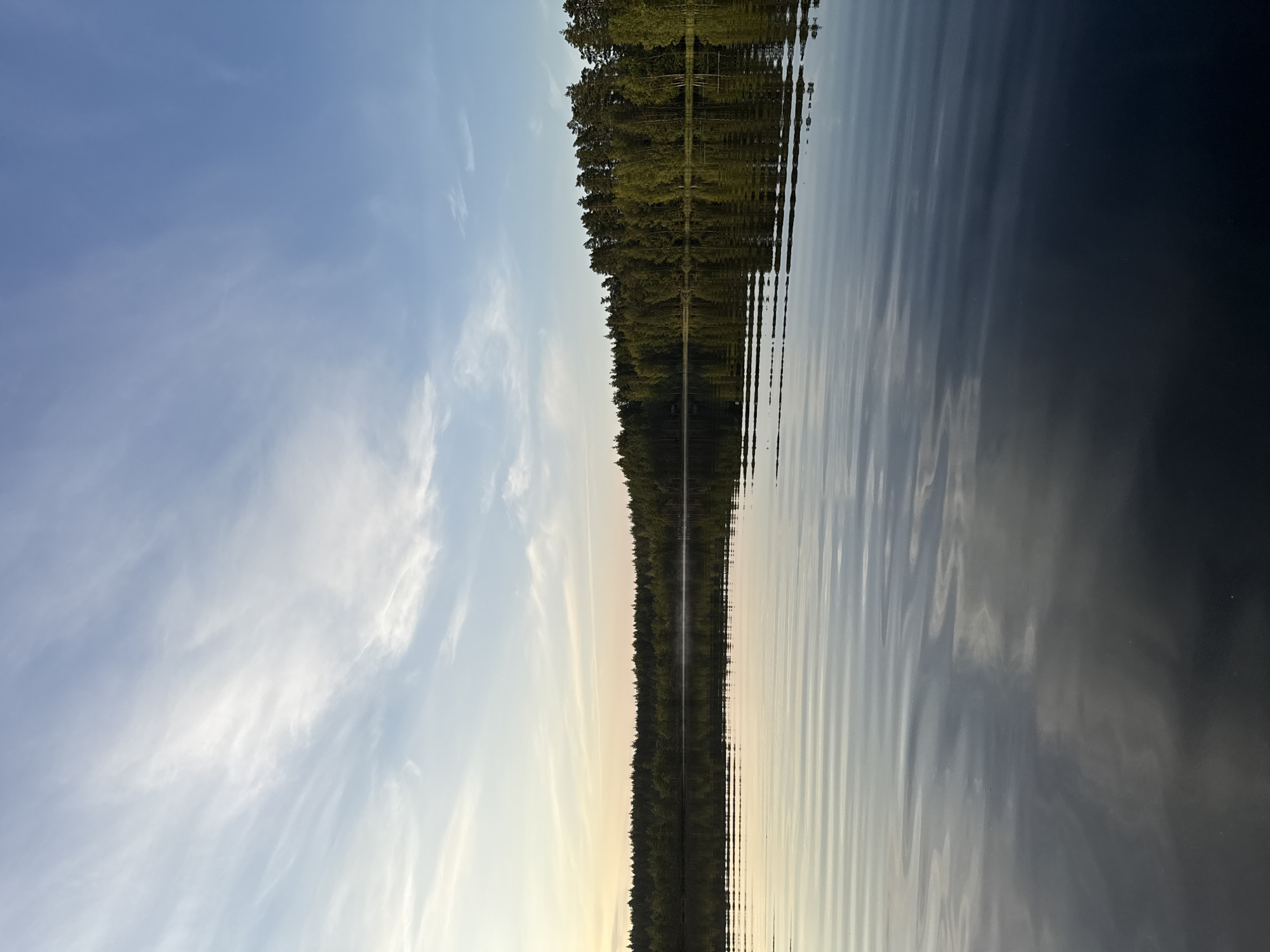 The image depicts a serene lake with a dense forest lining its far bank. The sky above is a soft blue with wispy white clouds, transitioning to a warm yellow hue near the horizon, suggesting either dawn or dusk. The lake's surface is calm, with gentle ripples reflecting the sky and trees. The scene appears to be a natural landscape, likely a remote or undeveloped area. Based on the provided context, this scene is located in the city of Kokemäki, Finland.