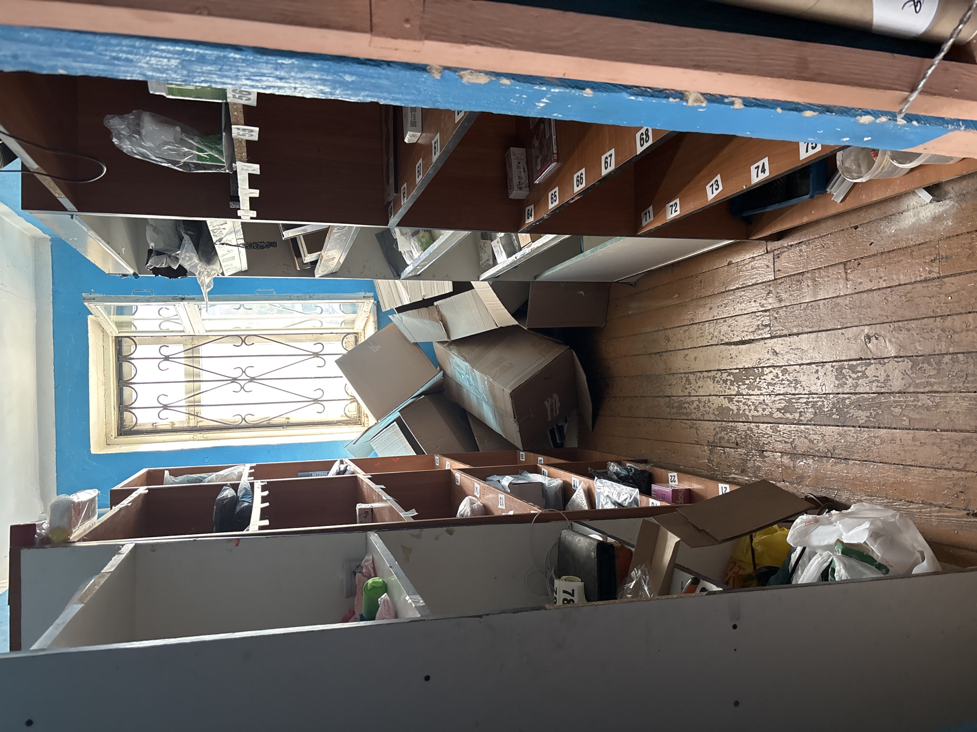 This media file shows a cluttered room with multiple shelves filled with various items. The shelves are wooden, with some appearing worn and others painted white. Many of the shelves have labels with numbers, such as 56, 60, 61, 64, 65, 66, 67, 68, 71, 72, 73, 74, and 21, 22. These numbered labels suggest the shelves might be part of a postal sorting facility, a storage system, or mailboxes.
The shelves are filled with a variety of objects, including cardboard boxes of different sizes, plastic bags containing items, and what appear to be rolls of paper or posters at the top of the shelves. Some shelves contain smaller, more organized items, while others are overflowing with disorganized clutter. In the foreground, there are also several large cardboard boxes piled up, some of which are open.
A window with a decorative metal grate is visible in the background, letting in natural light. The walls appear to be painted blue, and the floor is made of old wooden planks that show signs of wear and tear.
There are no people visible in the image. The scene appears to be indoors, possibly in a dimly lit area due to the presence of natural light from the window. The overall impression is one of disarray and accumulated items, suggesting a place that is either actively in use and being filled, or a storage area that is neglected. The location is identified as Vileyka, Belarus.