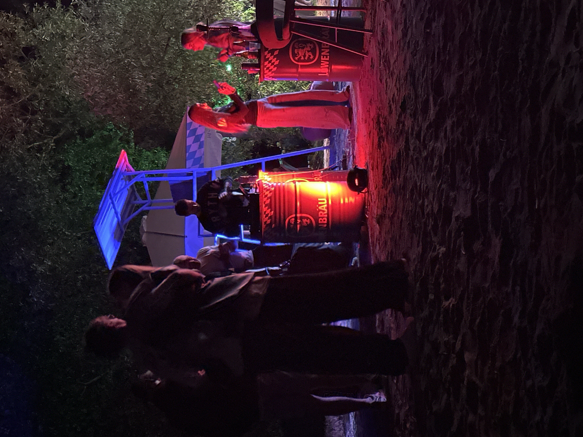 The image depicts an outdoor nighttime gathering taking place in Ждановіцкі сельскі Савет, Belarus, on an uneven, dark ground surface that appears to be sand or dirt. The scene is illuminated by artificial colored lights, primarily red and blue, creating distinct zones of light and shadow.
Approximately seven to nine individuals are present. In the left foreground and midground, a group of people are largely silhouetted, their facial features obscured by the ambient darkness. One individual in the immediate foreground is oriented with their back to the viewer.
At the center-right, a male individual stands behind a red metal barrel, illuminated by both red and blue light. He is operating what appears to be DJ equipment. He wears a dark top featuring the partial inscription "RYTHM." This barrel, which has "BRAU" visible (likely part of "LÖWENBRÄU"), functions as a support structure for the audio equipment. A dark, spherical object rests on the ground directly in front of this barrel.
To the right of the frame, two women are positioned near a second red metal barrel. This barrel is explicitly labeled "LÖWENBRÄU" and features a lion emblem; it serves as a high-top table with bottles and glasses visible on its surface. A tall stool is placed adjacent to it. The woman on the left has long, light-colored hair, wears a dark top and light-colored wide-leg pants, and is holding a mobile phone. The woman on the right, also with light-colored hair, wears a dark top and is holding an indistinct object in her hand. Both women are prominently illuminated by red light.
In the mid-background, a blue metal structure, resembling an elevated platform or lifeguard stand with a blue canopy, is visible. Behind this structure, a light-colored tent with a blue and white checkered pattern can be observed. Dark tree foliage forms the upper background, showing selective illumination from green, red, and blue lights. The ground in the immediate foreground and beneath the right barrel is strongly lit with red light, highlighting its irregular texture.