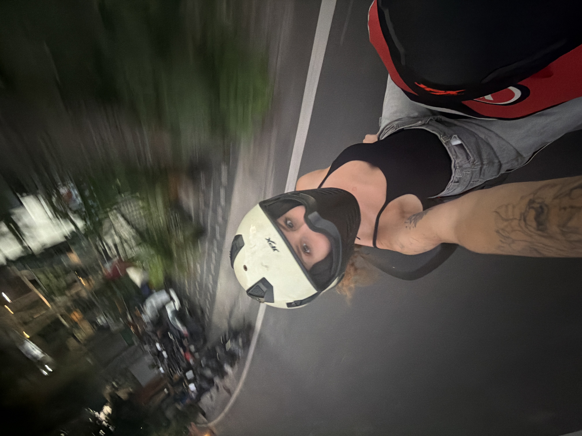 In this image, taken at night in the city of Mengwi, Indonesia, a woman is riding on the back of a motorcycle, looking towards the camera. She is wearing a white helmet with black accents and a black face covering. Her hair is reddish-blonde and visible beneath the helmet. She is dressed in a black tank top and light-wash denim overalls. Her left arm, visible in the foreground, is tattooed. The motorcycle's seat is black, and the background of the image is blurred, indicating movement. The background shows a street with parked motorcycles, buildings, and foliage, suggesting an urban or suburban setting. The lighting suggests it is nighttime, with artificial lights visible in the background.