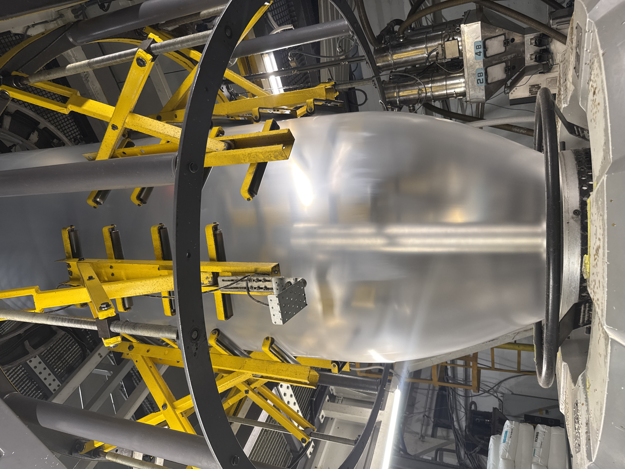 The image shows the interior of an industrial facility, likely in Perm, Russia, though this cannot be confirmed from the image alone. The main subject is a large, inflated, transparent plastic film tube, held in place by a complex network of yellow metal scaffolding and rollers. The scaffolding appears to be adjustable, with threaded rods and various connection points. Above the inflated film are several cylindrical metal components, possibly part of a heating or cooling system, along with associated wiring and tubing. A black circular frame partially surrounds the film, and a small silver metallic device is positioned near the film's surface. In the background, bags of materials are visible along the floor. The overall environment appears to be a manufacturing or processing plant.