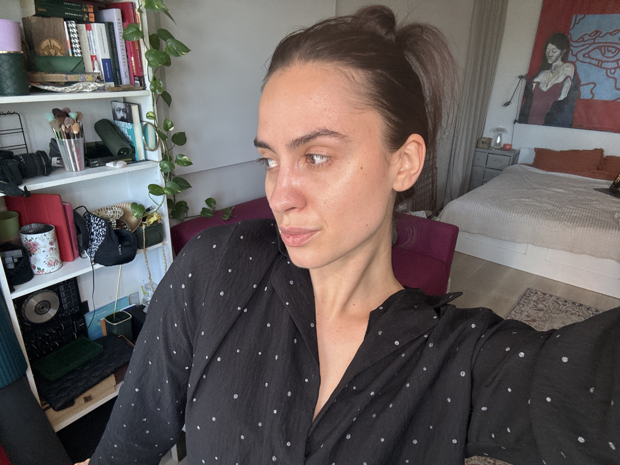 A woman with dark hair pulled back in a bun is the main subject. She is wearing a black blouse with white polka dots. She is looking off to the side with a neutral expression. Behind her is a bookshelf with books, a potted plant, and other miscellaneous items. A bed with orange pillows and a painting are visible in the background. The location in Krasnoyarsk, Russia is not discernible from the image.