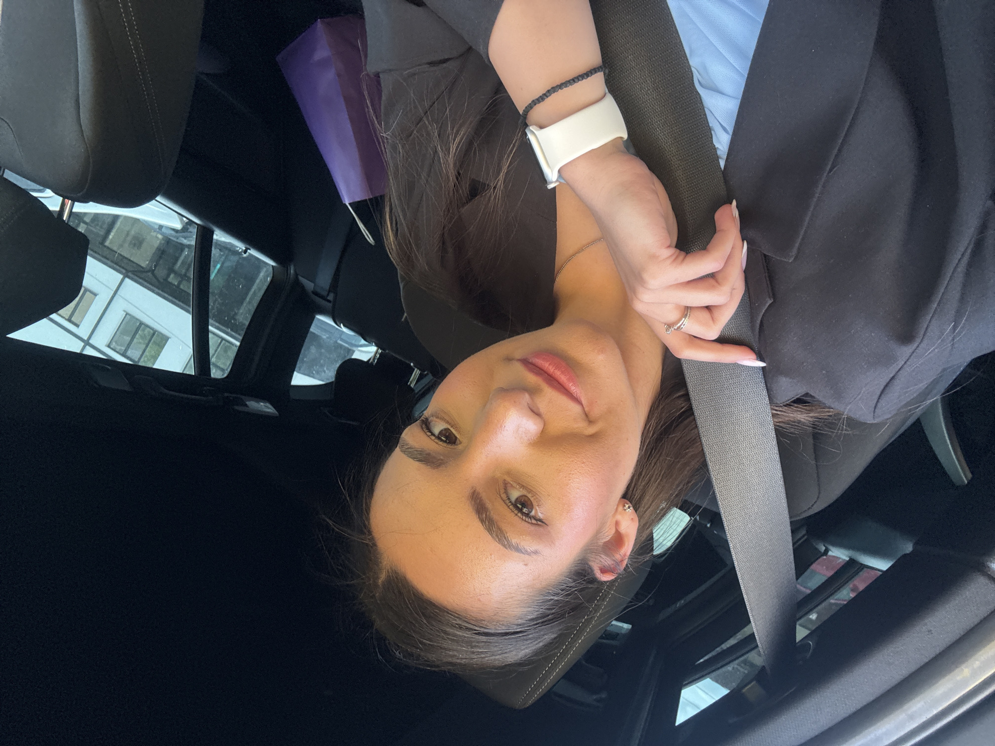 The image shows a woman in a car, presumably in Razgrad, Bulgaria (given the prompt's context), secured with a seatbelt. She is wearing a black blazer over a light blue shirt. A white smartwatch adorns her left wrist, accompanied by a black bracelet. A ring is visible on her finger. A purple shopping bag is partially visible in the background, hanging from the passenger seat headrest. The car's interior is primarily dark. Buildings are visible through the car windows.