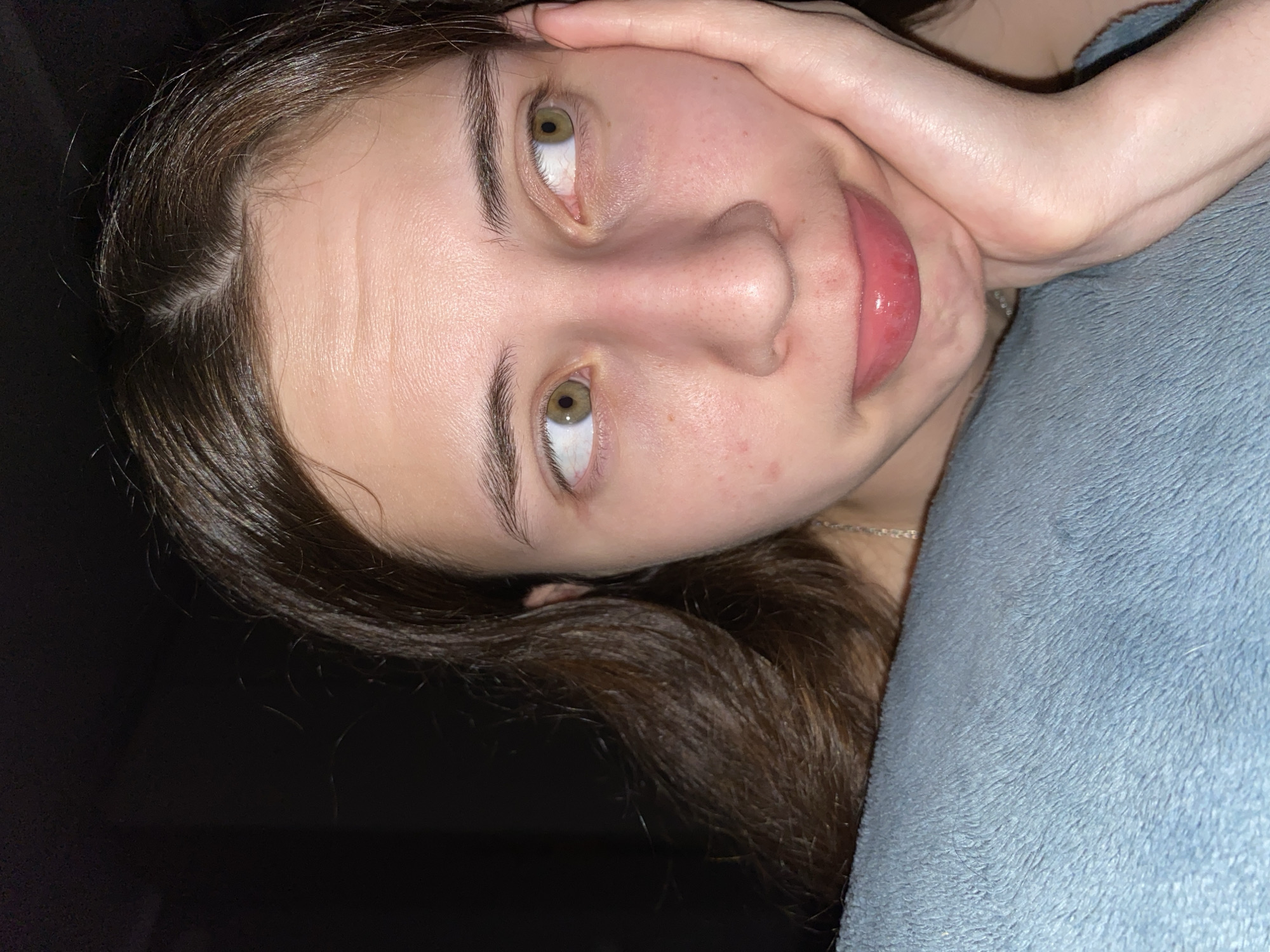 A close-up, angled headshot of a young woman. She is lying down on what appears to be a soft, blue blanket or cushion. Her brown hair is partially visible, with some strands falling towards the dark background in the bottom left corner. The woman's face is clear, revealing light skin with visible pores and minor skin imperfections. Her eyes are open, light green, and looking slightly upwards. Her hand is placed near her forehead. A thin silver necklace is visible. The location in the city of Votkinsk, Russia is not discernible from this image.