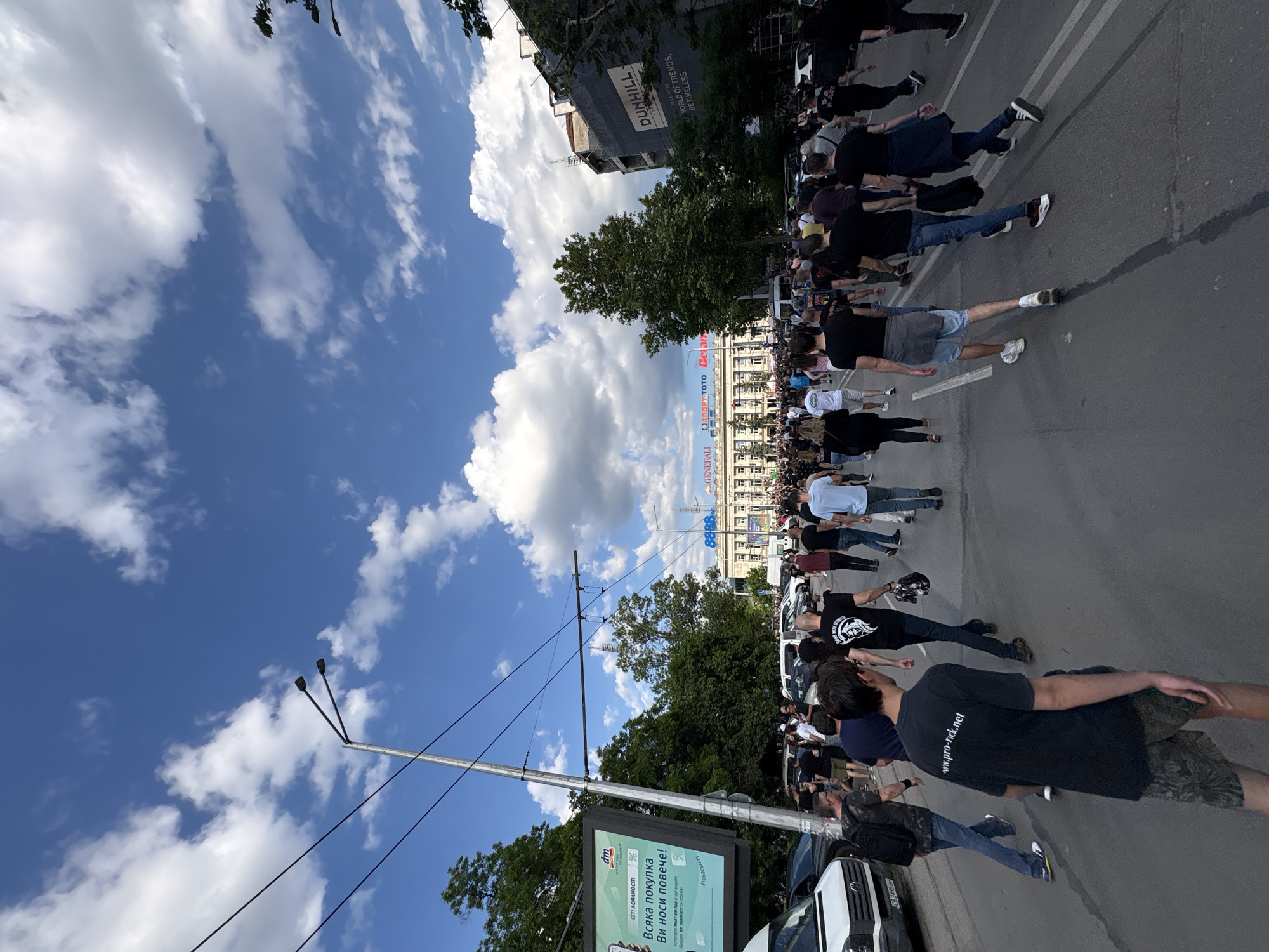 The image captures a street in Sofia, Bulgaria, where a long line of people are lying down on the asphalt. They are participating in what appears to be a demonstration or protest. The individuals, dressed in a variety of casual attire, are spread out along the street. Banners or flags hang overhead from buildings along the street. A billboard advertising "dm" and "Generali" are visible. Buildings bearing the name "Dunhill" appear in the background. The sky is blue with scattered clouds. Cars are parked on the side of the street.