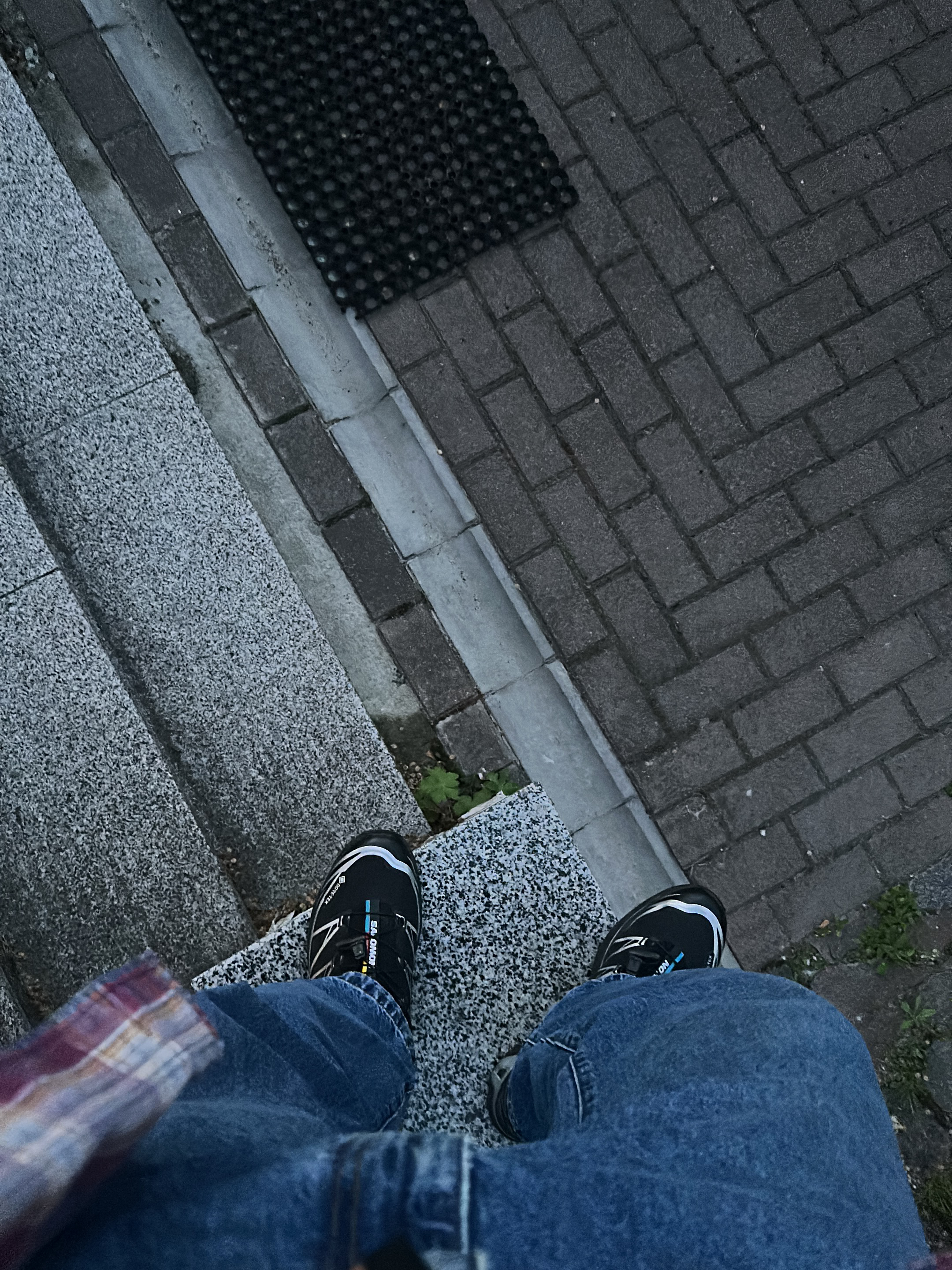The image shows a first-person perspective of someone standing on the top step of a set of concrete stairs. The person is wearing blue jeans and black and blue athletic shoes. To the right of the stairs is a brick walkway. At the top of the brick walkway is a black rubber mat. Based on the provided information, the location is Kriukivshchyna, Ukraine.