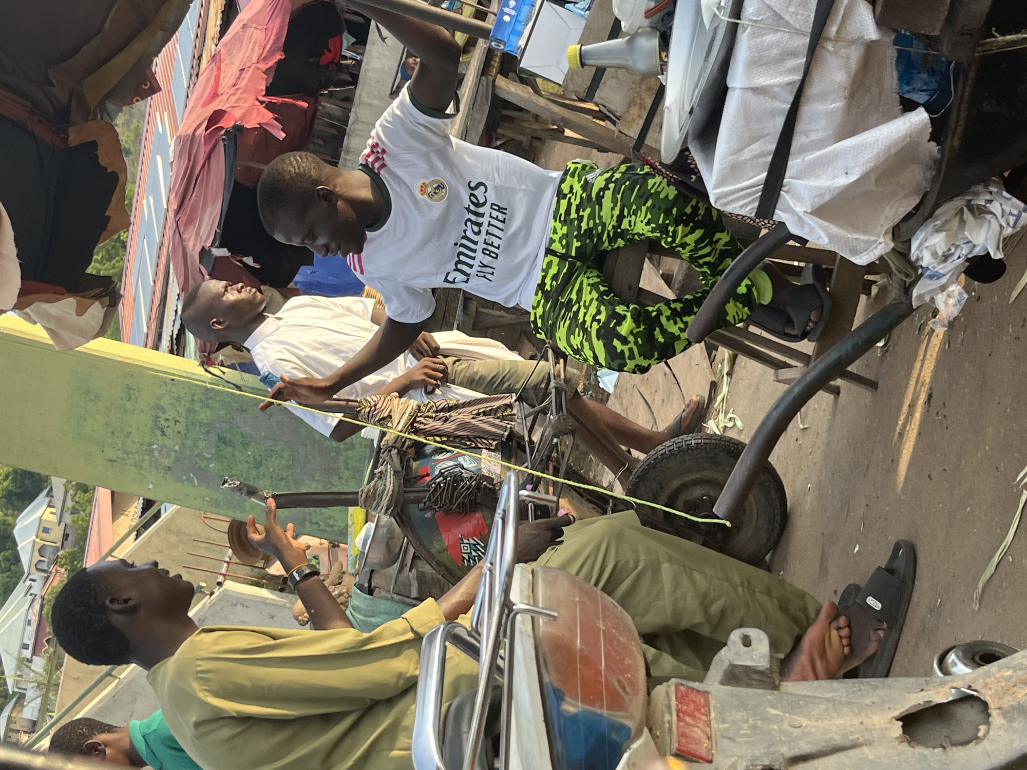 In a bustling outdoor market, possibly in a city, a group of individuals are engaged in conversation. The scene is lit by natural sunlight, casting shadows and highlighting the textures of the environment. In the foreground, a person wearing olive green trousers and a matching shirt is seated on a motorcycle. Their left leg is extended forward, with a black sandal on their foot. To the right, another person is seated, wearing a white t-shirt with "Emirates Fly Better" printed on it and brightly colored camouflage trousers. They are gesturing with their hands, appearing to be animated in their conversation. Behind them, another person in a white shirt stands, facing the seated individual. In the background, tents and stalls suggest a marketplace setting, with buildings and foliage visible further in the distance. The overall impression is one of everyday life in a vibrant community setting.