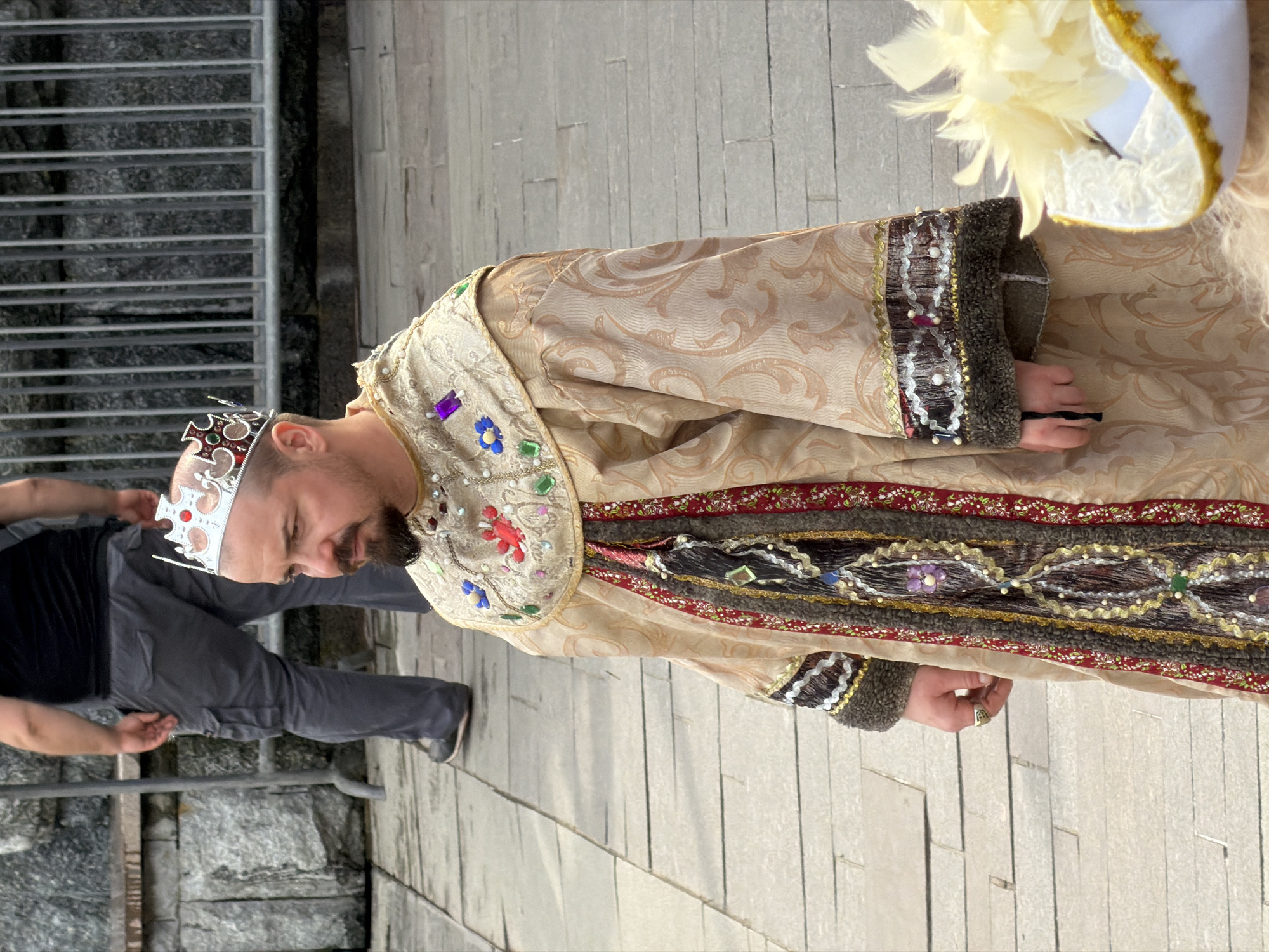 The image shows a man dressed in an ornate, possibly historical or theatrical costume, including a crown, which suggests a king or royal figure. The costume is detailed with embroidery, gemstones, and fur trim. He appears to be standing on a paved surface with a metallic railing visible to his left. Another person is partially visible to his left. The location cannot be reliably determined as Moscow, Russia based on the visual information provided in the image.