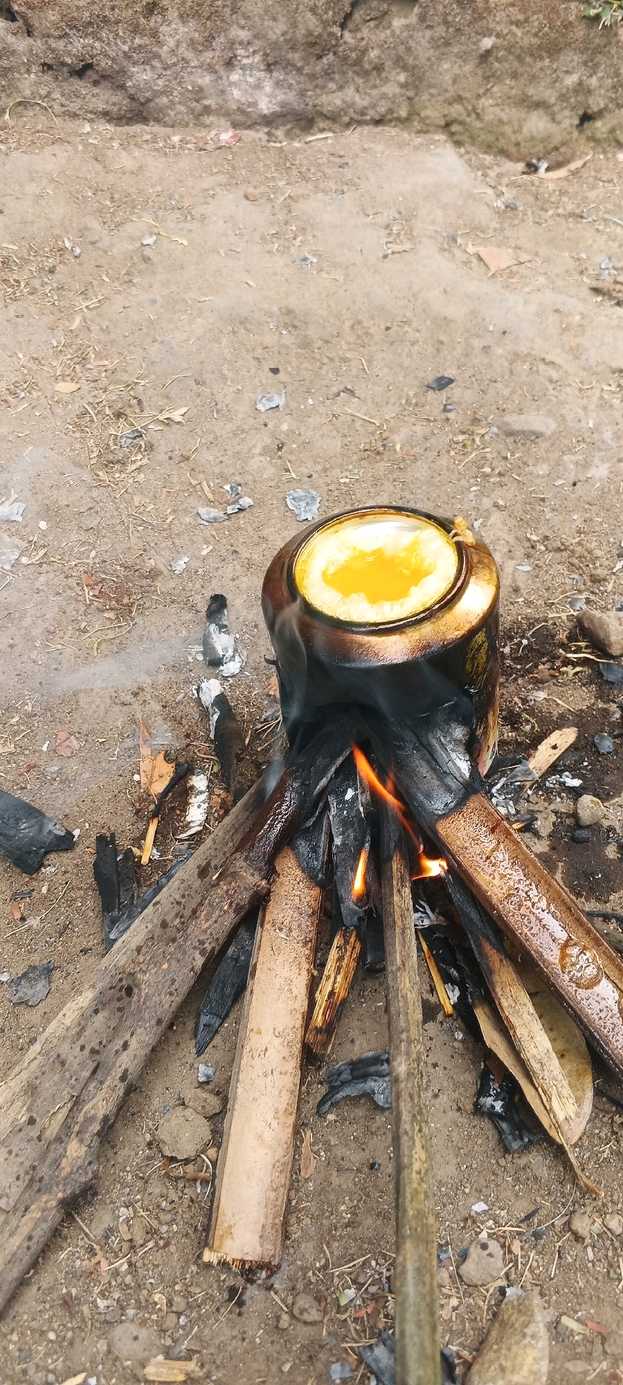 A rustic cooking setup is depicted in an outdoor, earthy environment, likely in the vicinity of Penanggungan, Indonesia. A metallic pot, blackened by soot, is perched precariously atop a makeshift fire constructed from wood and charcoal. Flames are visible licking at the base of the pot, which contains a bright yellow, semi-liquid substance, possibly egg yolk or a similar culinary preparation. Smoke drifts upwards from the fire, adding to the scene's primitive atmosphere. The ground is a mixture of soil, ash, small stones, and dry vegetation, consistent with an outdoor, undeveloped setting. The overall impression is one of resourcefulness and traditional cooking methods.