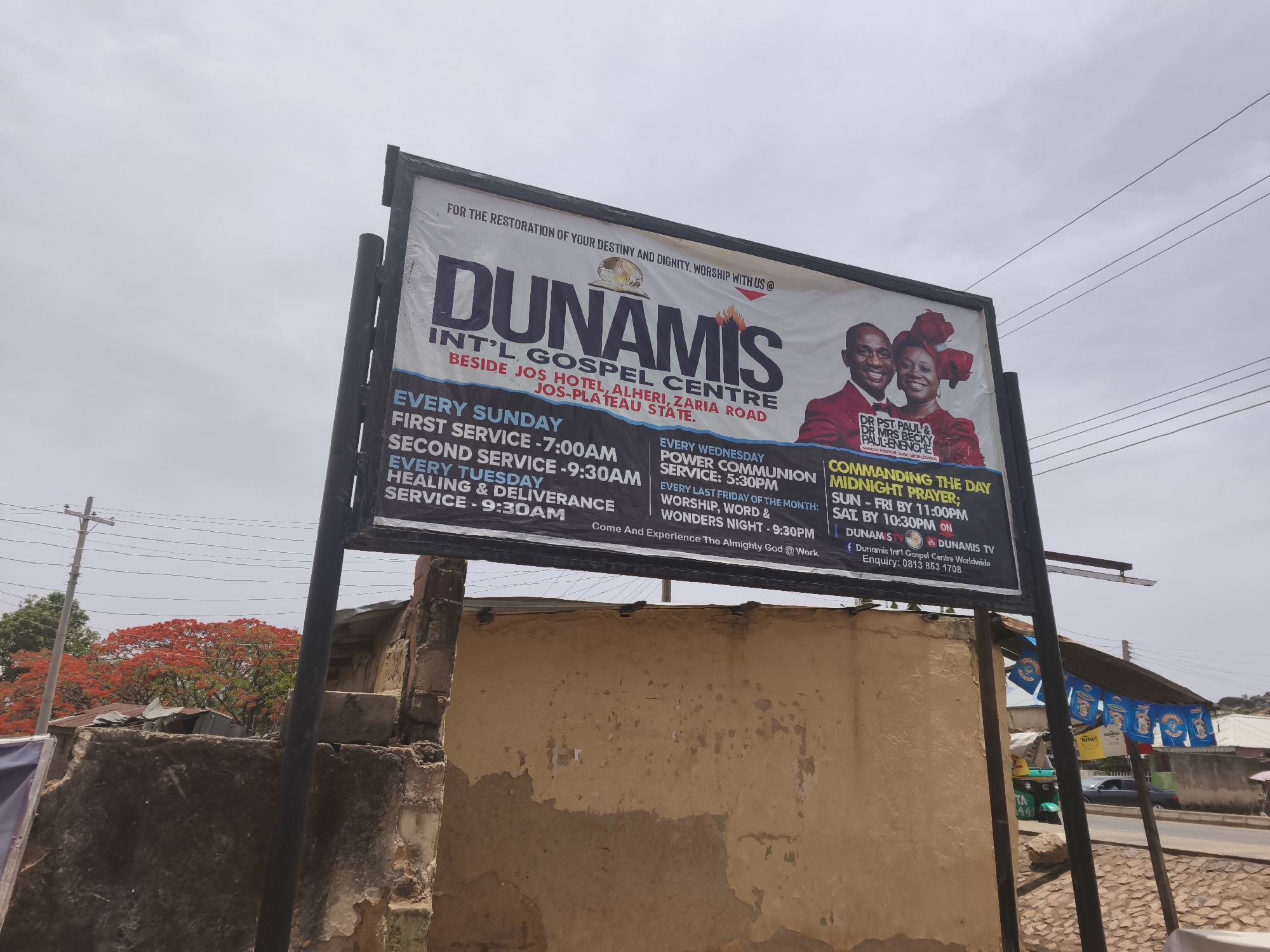 A large, rectangular signboard for "Dunamis Int'l Gospel Centre" dominates the center of the frame, set against an overcast sky during daytime. The sign features the church's name in bold lettering, along with its address: "BESIDE JOS HOTEL, ALHERI, ZARIA ROAD, JOS-PLATEAU STATE."
The signboard advertises various services and events:
* **Every Sunday:** First Service - 7:00 AM, Second Service - 9:30 AM
* **Every Tuesday:** Healing & Deliverance Service - 9:30 AM
* **Every Wednesday:** Power Communion Service - 5:30 PM
* **Every Last Friday of the Month:** Worship, Word & Wonders Night - 9:30 PM
* **Commanding The Day Midnight Prayer:** Sun-Fri by 11:00 PM, Sat by 10:30 PM
It also displays smiling portraits of "DR PST PAUL & DR MRS BECKY PAUL-ENENCHE," identified as Senior Pastors, DIGC Worldwide. The slogan "FOR THE RESTORATION OF YOUR DESTINY AND DIGNITY, WORSHIP WITH US @" and "Come And Experience The Almighty God @Work" are visible. Contact information including "DUNAMIS STV," "DUNAMIS TV," a Facebook handle, and an enquiry phone number (0813 853 1708) are listed.
The setting appears to be a street in New Stadium, Nigeria. Behind the signboard, a dilapidated building with peeling plaster is visible. Further back, a vibrant red-blossomed tree (possibly a Flamboyant tree), utility poles, and power lines extend across the frame. In the distance to the right, more buildings, a vehicle, and what appear to be hanging bags labeled "NIDO" and "CONSEG" suggest a commercial or residential area. The overall weather is cloudy, contributing to a soft, diffused light.