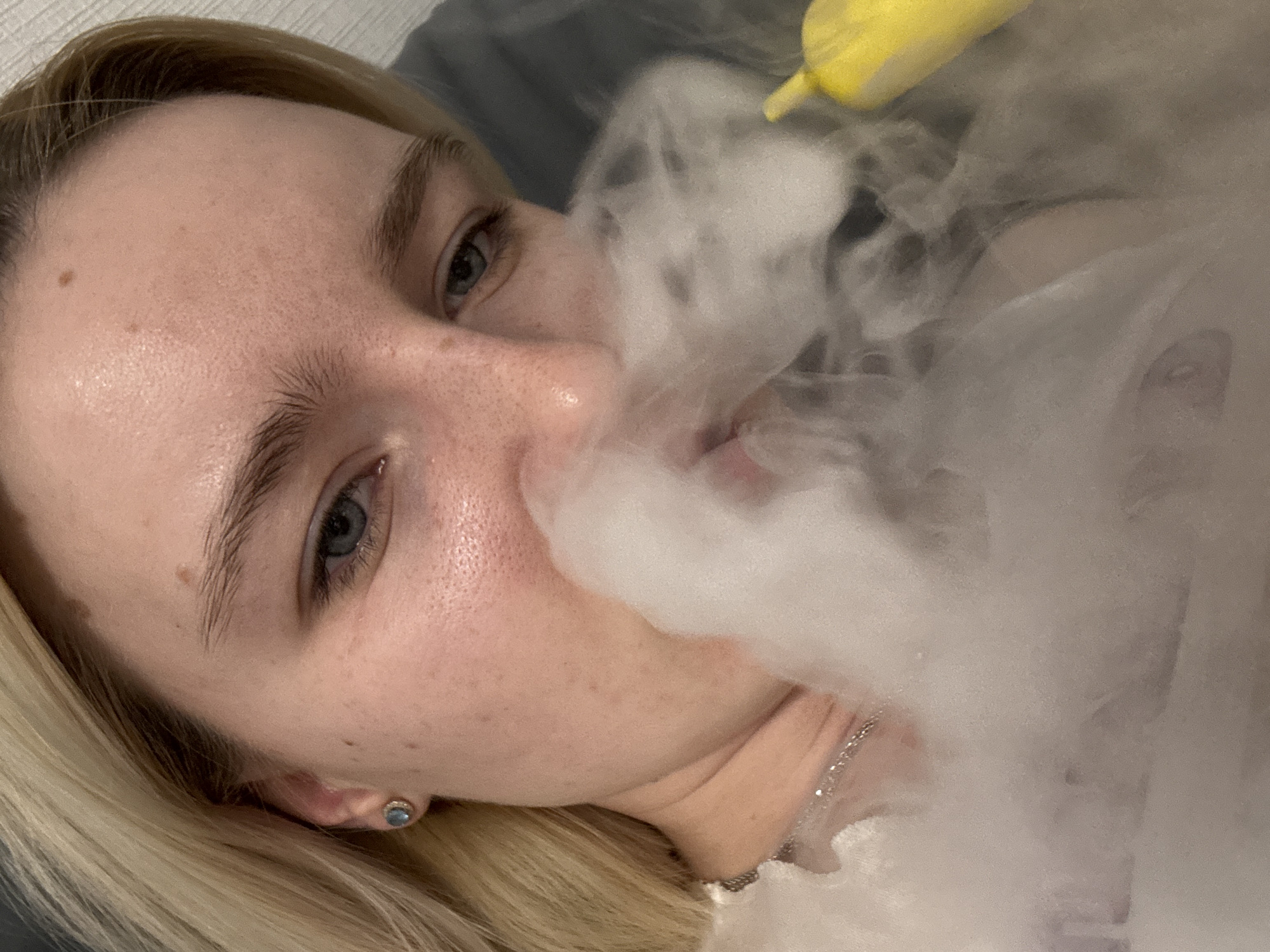 A close-up, angled shot shows a blonde woman with blue eyes surrounded by white smoke. Her face is visible from her forehead down to her neck. A small yellow object is partially visible above the smoke. The woman is wearing a small earring and a delicate necklace. Her expression is obscured by the smoke. No information regarding a location is available.