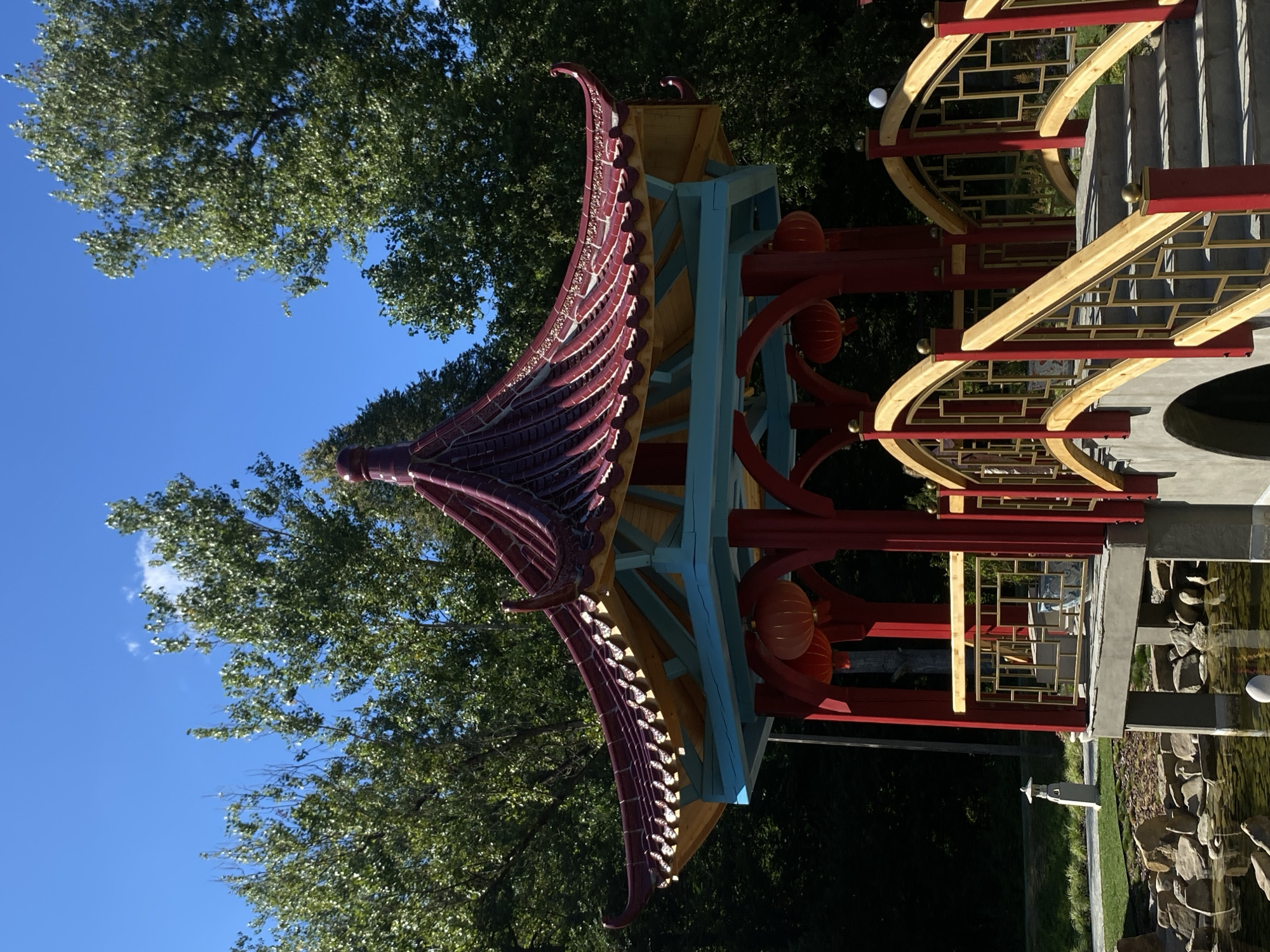 The image captures a vibrantly colored Chinese-style pagoda structure set amidst lush greenery and a bright blue sky. The pagoda features a multi-tiered, upward-curving roof in shades of deep red and yellow, with intricate decorative elements along its edges. The supporting pillars are painted red, and a light blue ceiling is visible beneath the roof. Two orange lanterns hang from the underside of the pagoda.
In the foreground, a decorative bridge with red pillars and golden latticework railings leads up to the pagoda. The bridge spans over a pond with visible rocks and clear, rippling water. To the left of the bridge, a small, white, lantern-shaped garden ornament stands on a grassy area. The background is dominated by dense, green trees, indicating a natural or park-like setting. The overall scene suggests a tranquil and aesthetically pleasing environment, likely a garden or a cultural park.
The described pagoda and surrounding garden area are part of the "Ogród Sensoryczny" (Sensory Garden) located in Muszyna, Poland. This garden is known for its diverse plantings, water features, and various architectural elements designed to stimulate the senses.