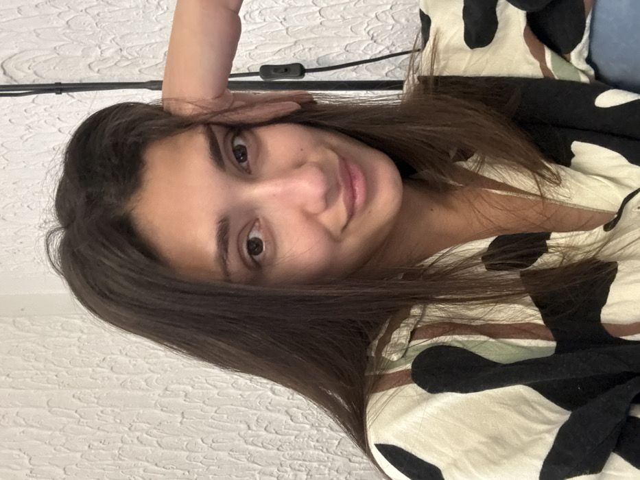 A forensic examination of this image, taken in Zankovići, Montenegro, reveals a single female subject positioned indoors. The subject is depicted from the mid-chest up, featuring medium-dark brown hair, brown eyes, and a fair to medium skin tone. Her expression includes a slight, closed-lip smile. Her left hand is placed against the left side of her face, with fingers partially visible. She wears a collared shirt characterized by an irregular pattern of black and brown shapes on a light beige base. The immediate background is a white wall with a textured, stucco-like finish. A black electrical cord, equipped with an in-line switch, is present vertically along the right edge of the frame against the wall.
