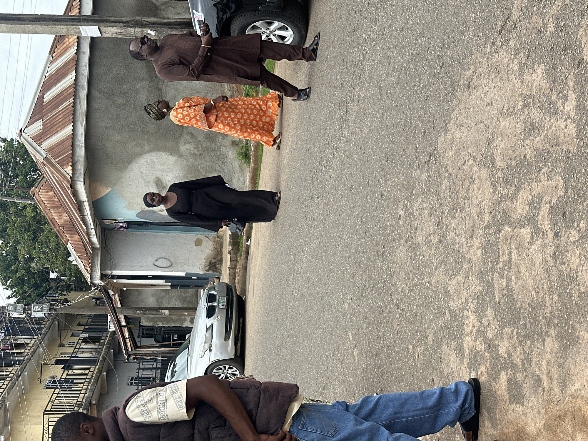 This image captures a street scene in Unknown, Unknown, under an overcast sky. Three individuals are walking from left to right across the mid-ground. The leftmost walker is a woman dressed in a long black gown, black head covering, and dark sunglasses, carrying a black handbag. To her right, a second woman wears a vibrant orange patterned dress with a patterned headwrap and gold-colored flat sandals. The third walker, a man on the far right, is dressed in a brown traditional tunic and trousers, dark sunglasses, and black slide sandals, holding a smartphone.
In the foreground, partially visible on the left, is another person wearing a dark brown sleeveless vest over a light t-shirt with "SOLD OUT" repeatedly printed, blue jeans, and dark slide sandals.
In the background, a silver BMW sedan with license plate "GWA-443BR" is parked on the left side of the road, and a portion of a black SUV is visible on the far right behind the man in brown. Buildings line the background: a multi-story, light-colored building with dark metal balconies on the left, and a smaller, single-story building with concrete walls and a prominent, rusted corrugated metal roof on the right. A utility pole with numerous wires stands near the center of the background. The asphalt road surface shows wear and patches of lighter discoloration.